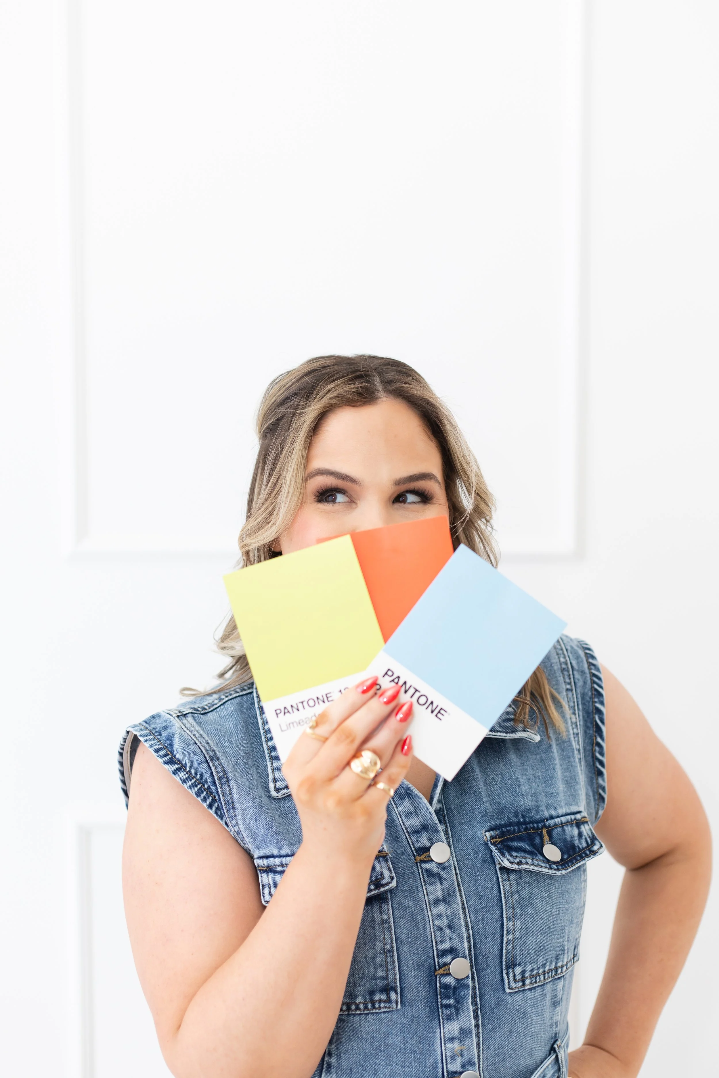 A woman with blonde hair wearing a sleeveless denim jacket holds up a Pantone color guide with colors including yellow, orange, and light blue, standing in front of a plain white background.