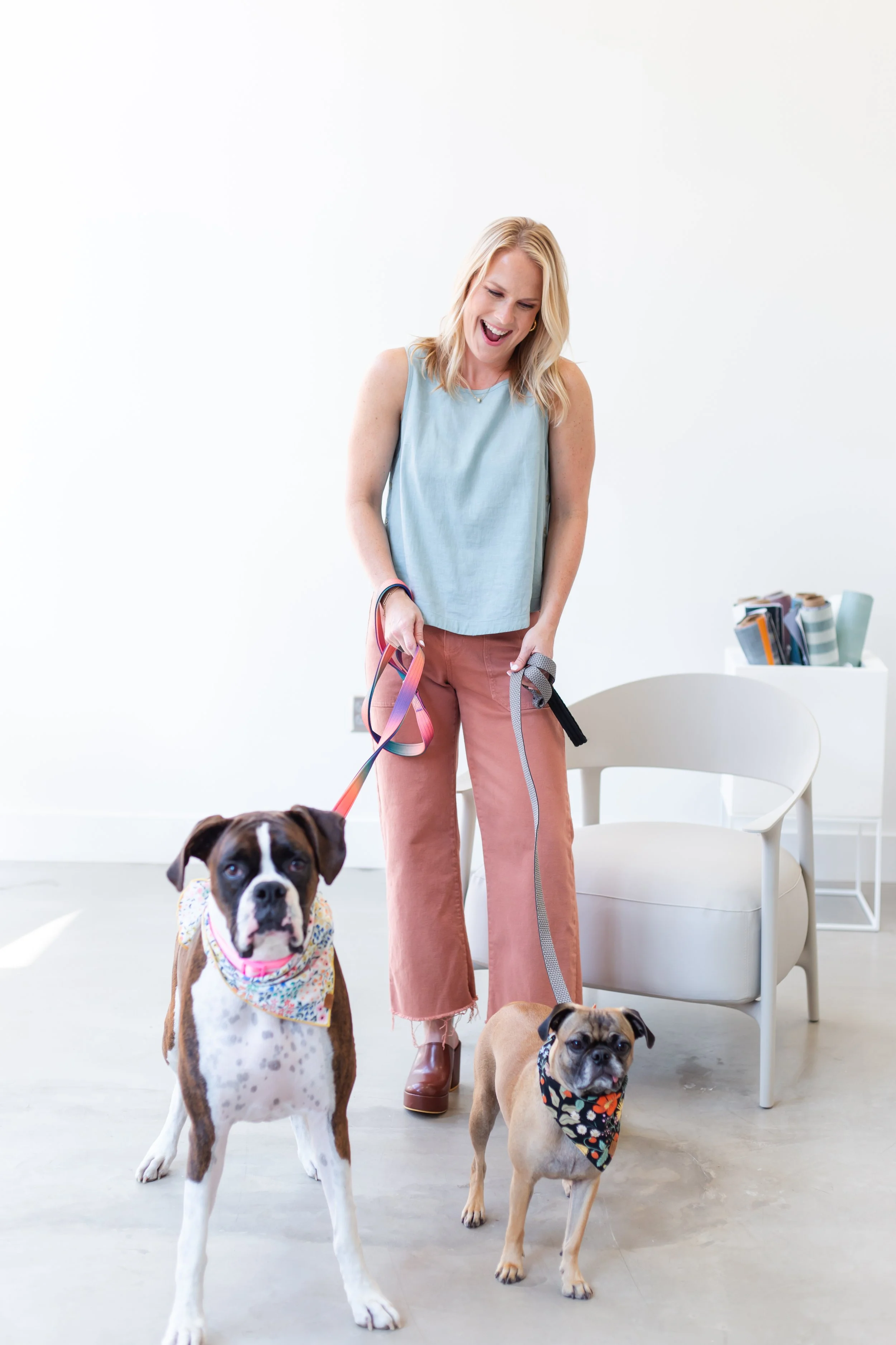 A young woman with blonde hair wearing a light blue sleeveless top and pink pants, smiling and holding the leashes of three dogs inside a bright, minimalist room.