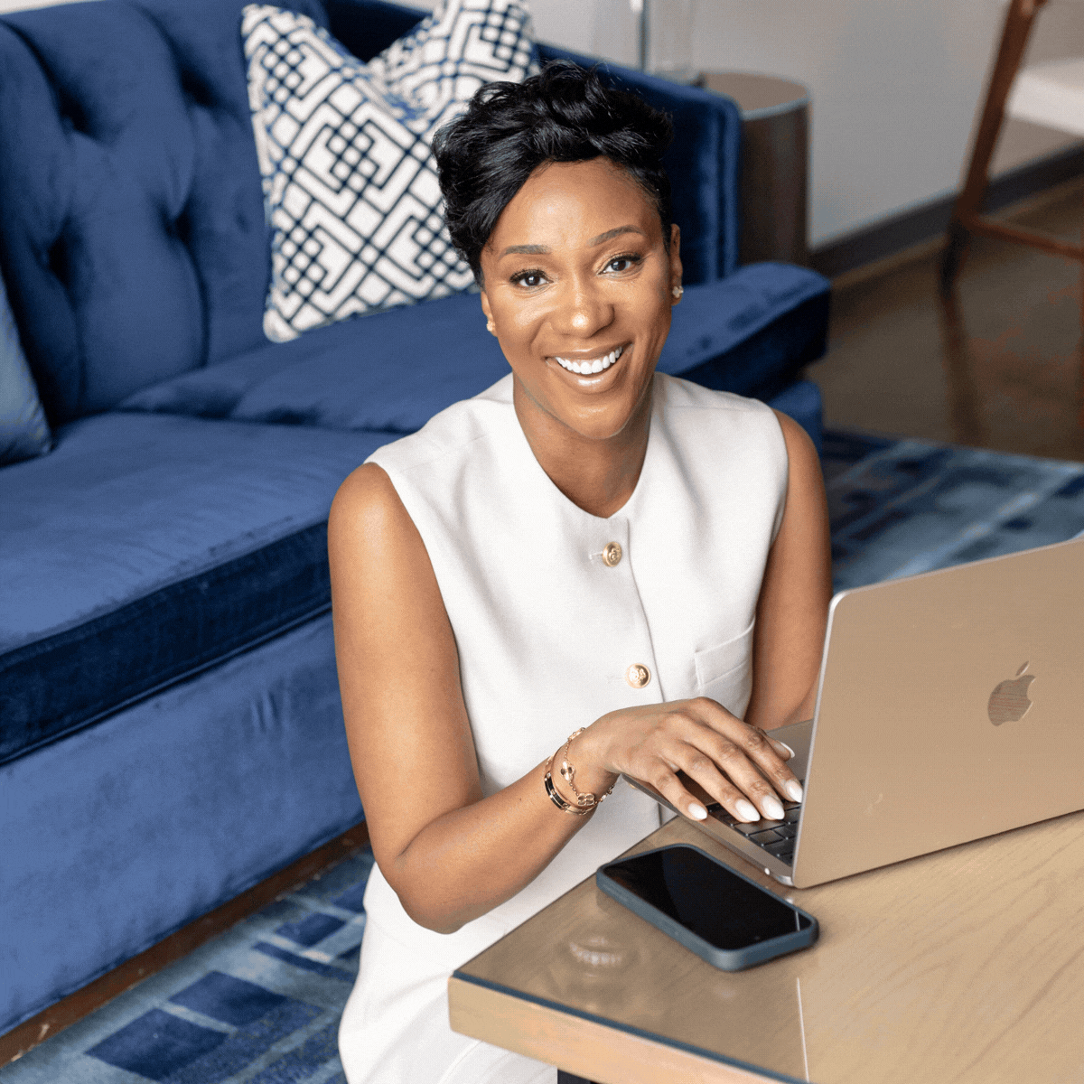 A woman with short black hair and a sleeveless beige top smiling and working on a silver MacBook at a wooden table, with a smartphone nearby, in a living room with a blue couch and patterned pillows.