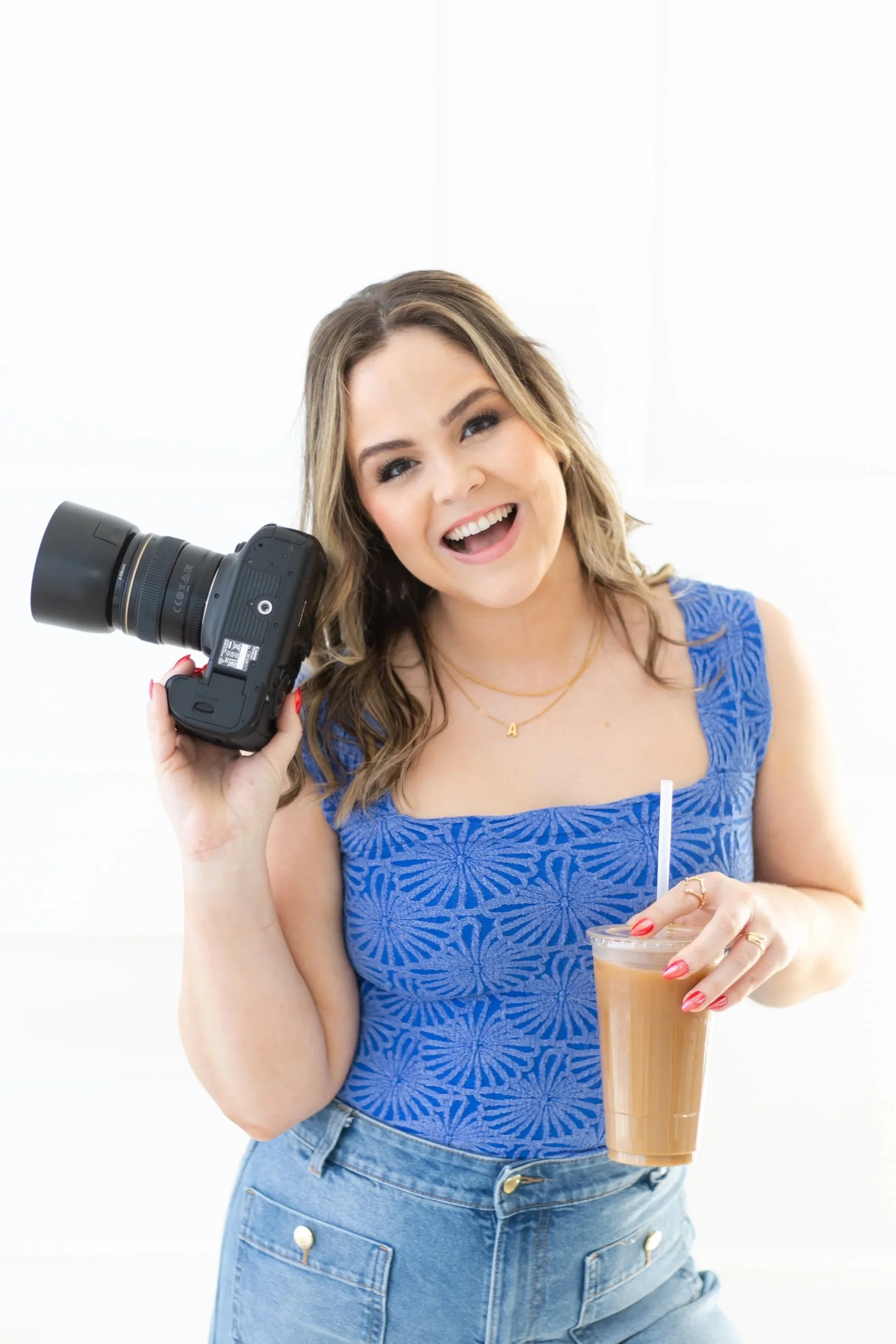 Smiling woman with light brown hair and blue eyes holding a camera in one hand and a cup of iced coffee with a straw in the other, wearing a blue lace top and denim shorts.