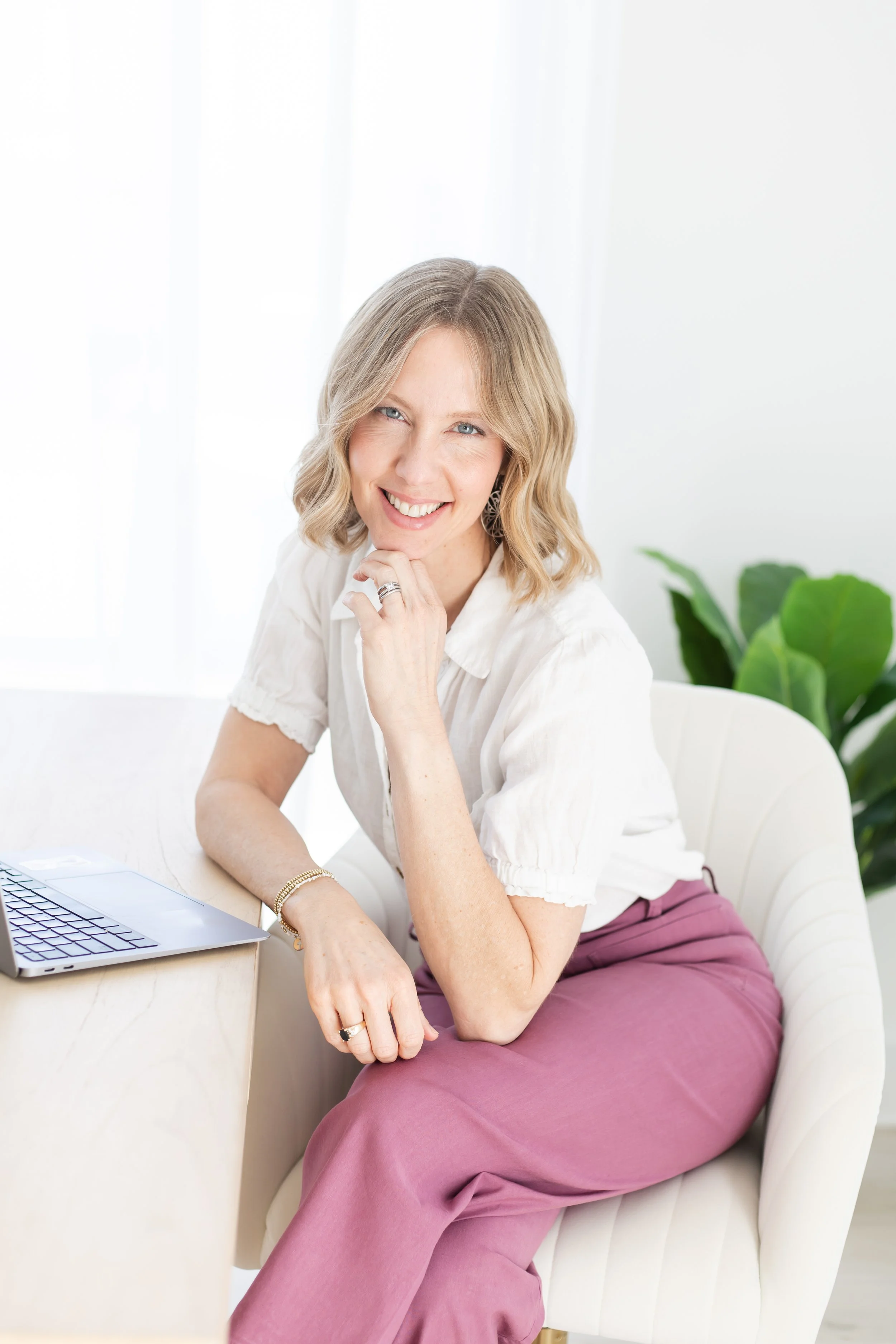 A woman with blonde wavy hair, wearing a white blouse and mauve pants, is sitting on a white sofa in a bright room with white walls, a large window, and a green plant in the background. She is smiling and resting her chin on her hand, with a laptop and some papers on the table next to her.