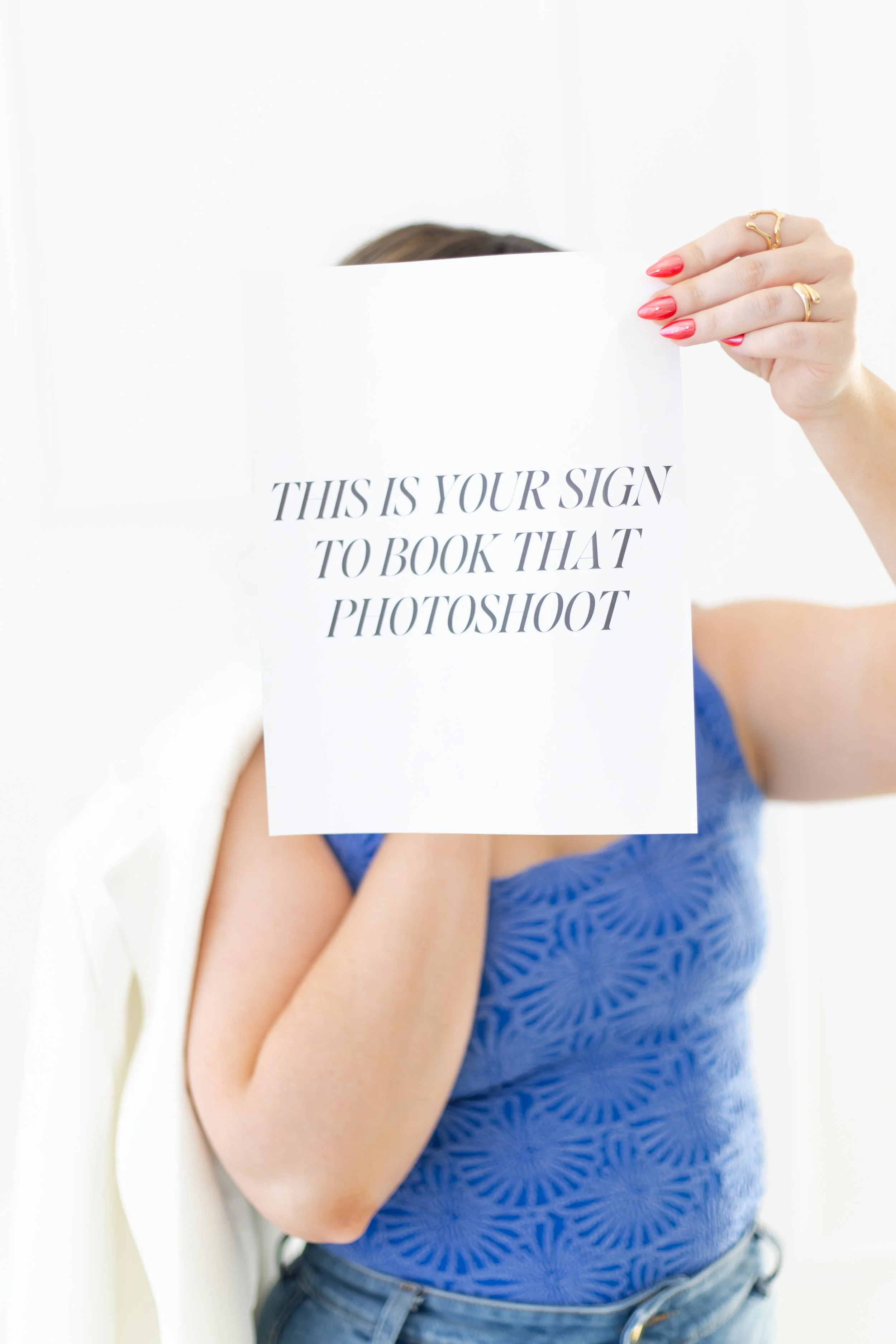 Person holding a sign that reads 'This is your sign to book that photoshoot'; the person has red nails and wears rings, a blue top with a circular pattern, and light blue jeans, with a white chair partially visible in the background.