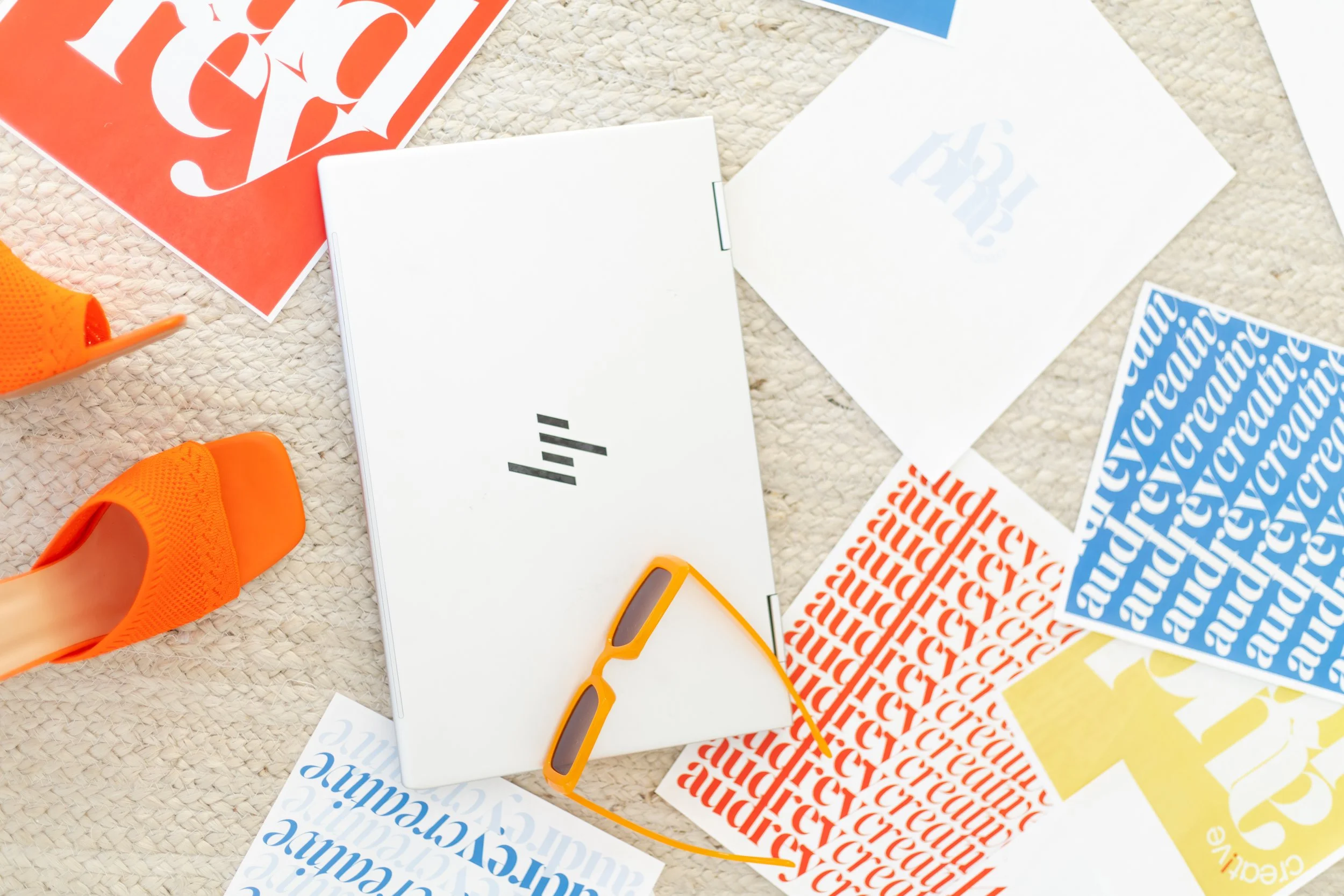 A flat lay of various colorful stickers, papers, and crafting supplies on a beige textured surface, including orange sneakers, orange sunglasses, and assorted printed sheets with cursive and block text in different colors.