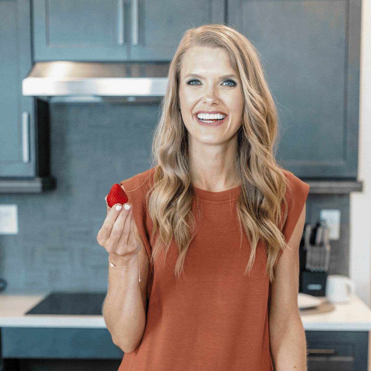 A smiling woman with long wavy blonde hair holds a strawberry in her right hand while standing in a modern kitchen.