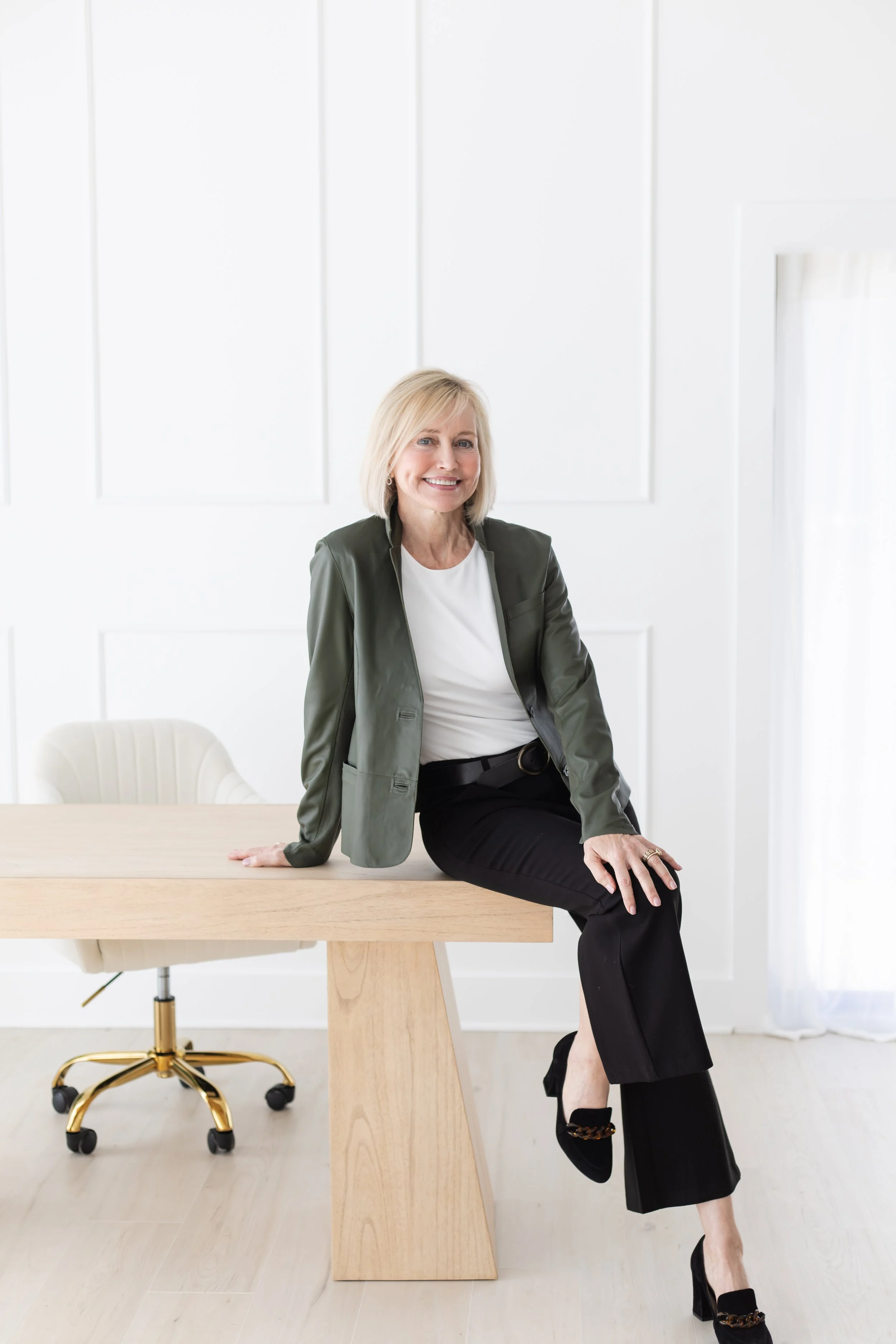 A woman with blonde hair, wearing a white shirt, dark pants, and a green jacket, is sitting on the edge of a wooden table in a bright, minimalistic room with white walls and curtains.