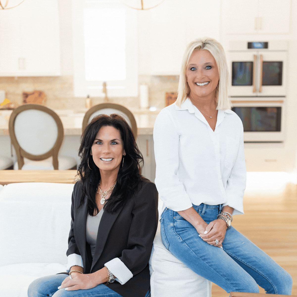 Two women sitting on a kitchen island in a bright, modern kitchen, smiling at the camera.
