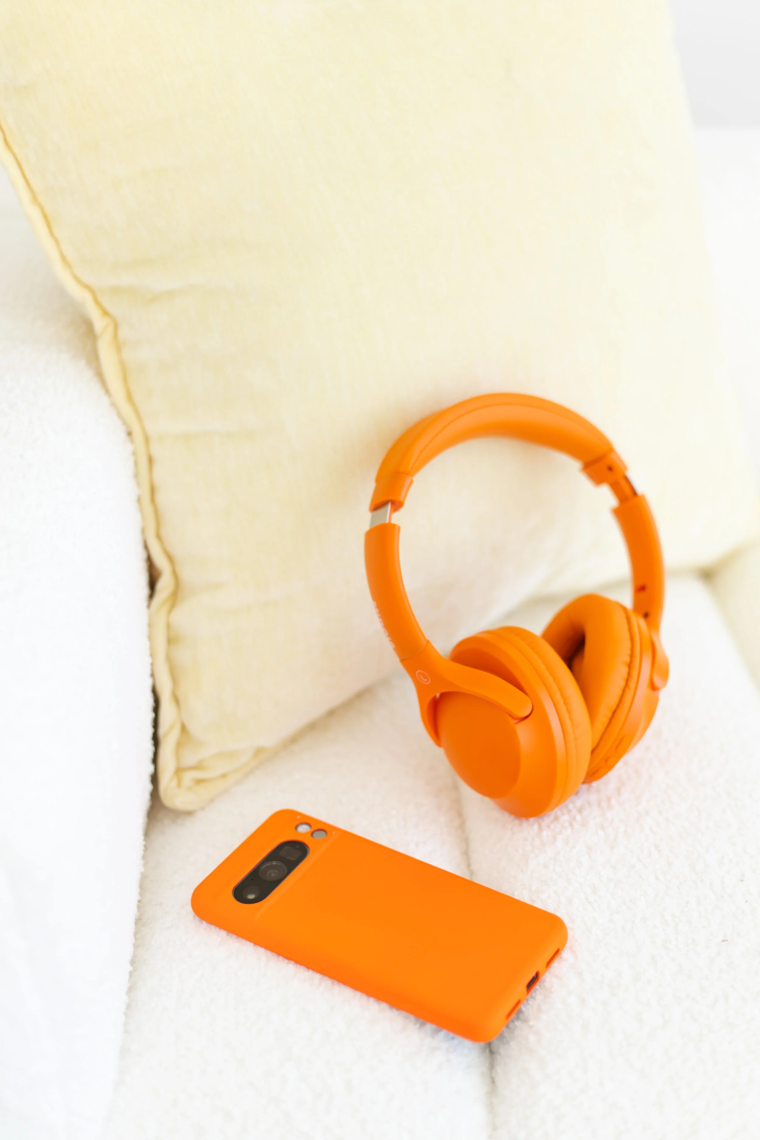 An orange smartphone, orange headphones, and a yellow pillow on a white textured surface.