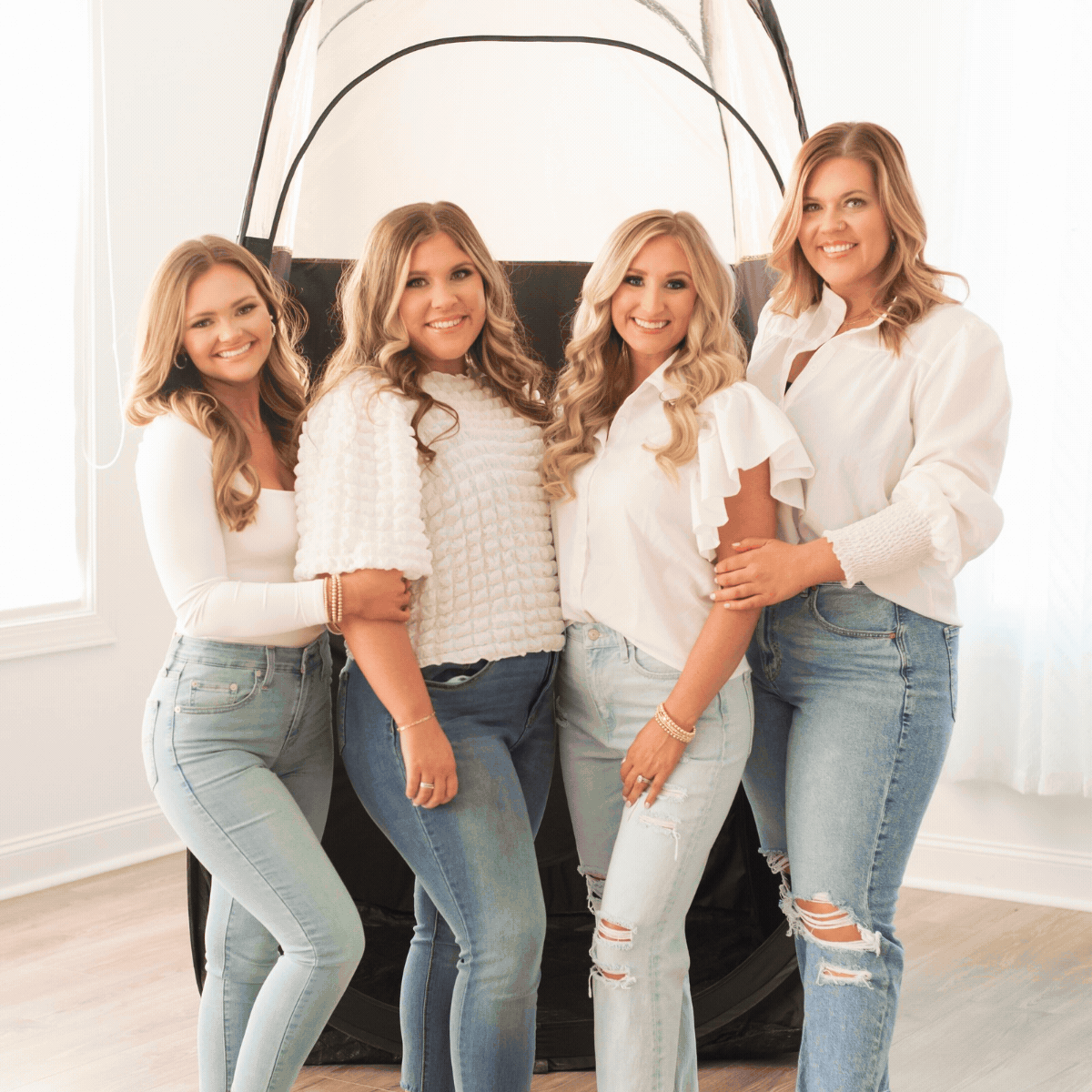 Four women with blonde hair wearing white tops and jeans, smiling and standing together in a bright room with a photography backdrop.