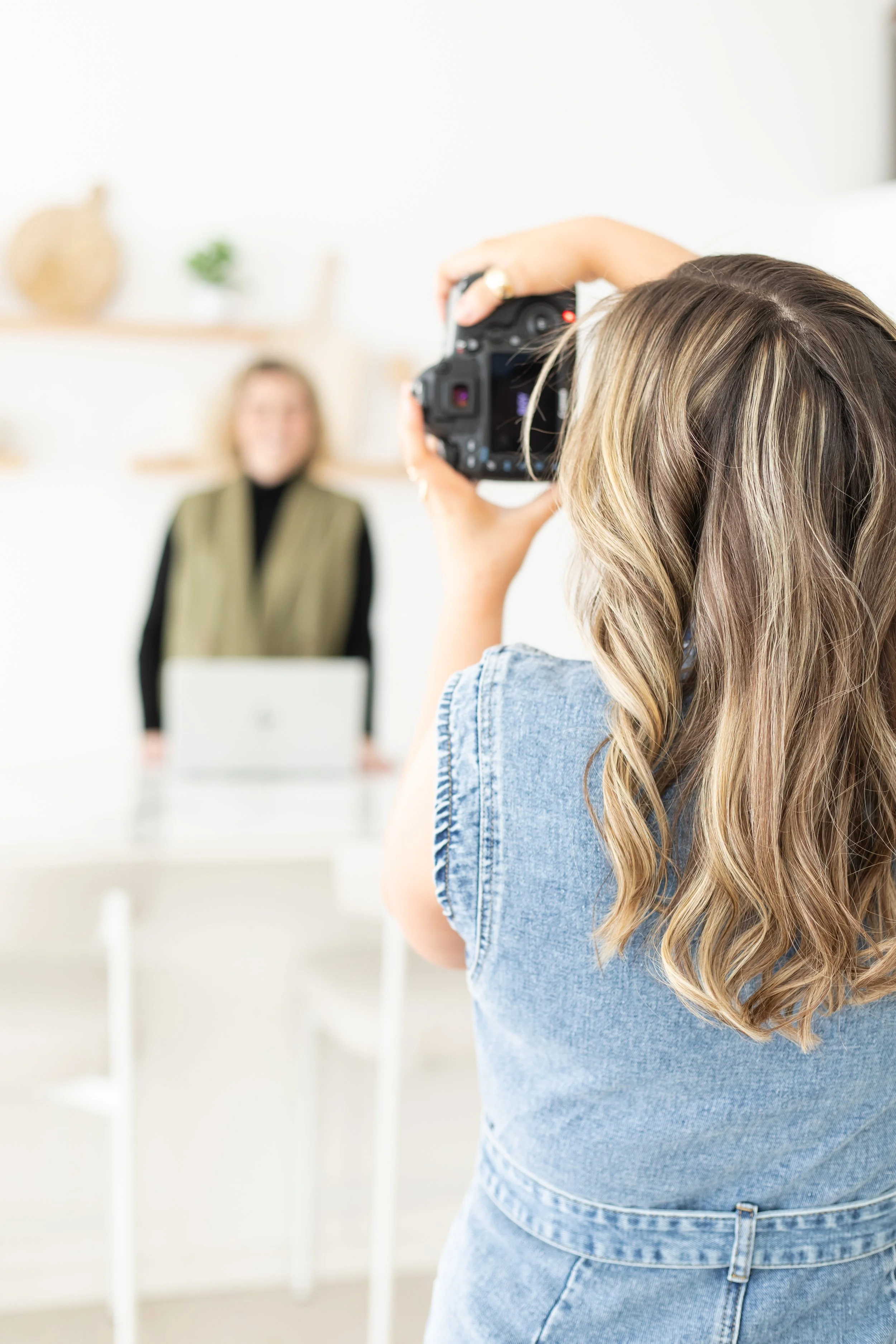 Person taking a photograph of a woman with blonde hair standing in a bright, minimalistic room with white walls and wooden shelves.