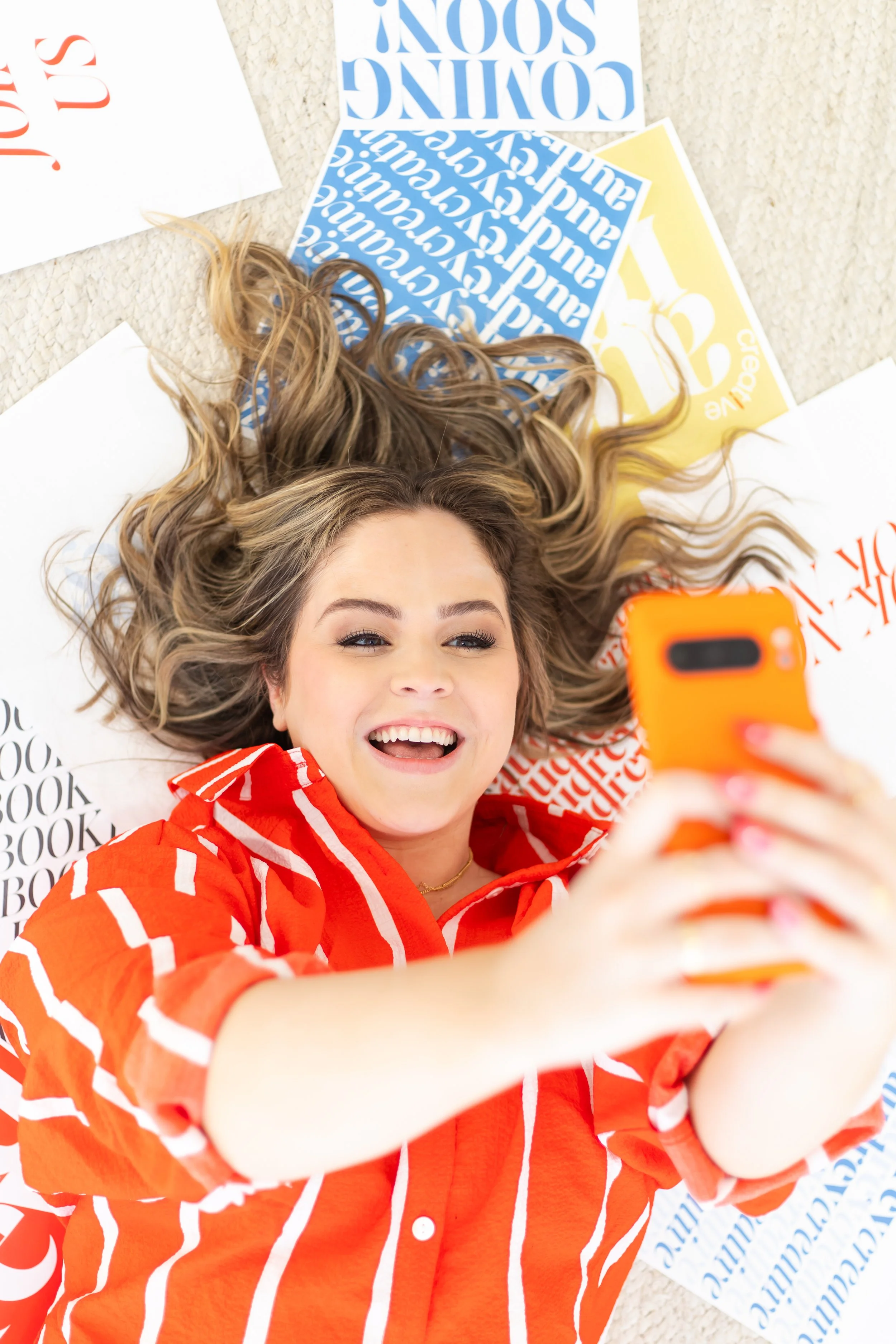 A young woman with long, wavy blonde hair lying on a carpeted floor, taking a selfie. She is smiling and wearing a bright red shirt with white vertical stripes. Several printed sheets of paper with text and graphics are spread around her, some partially visible.