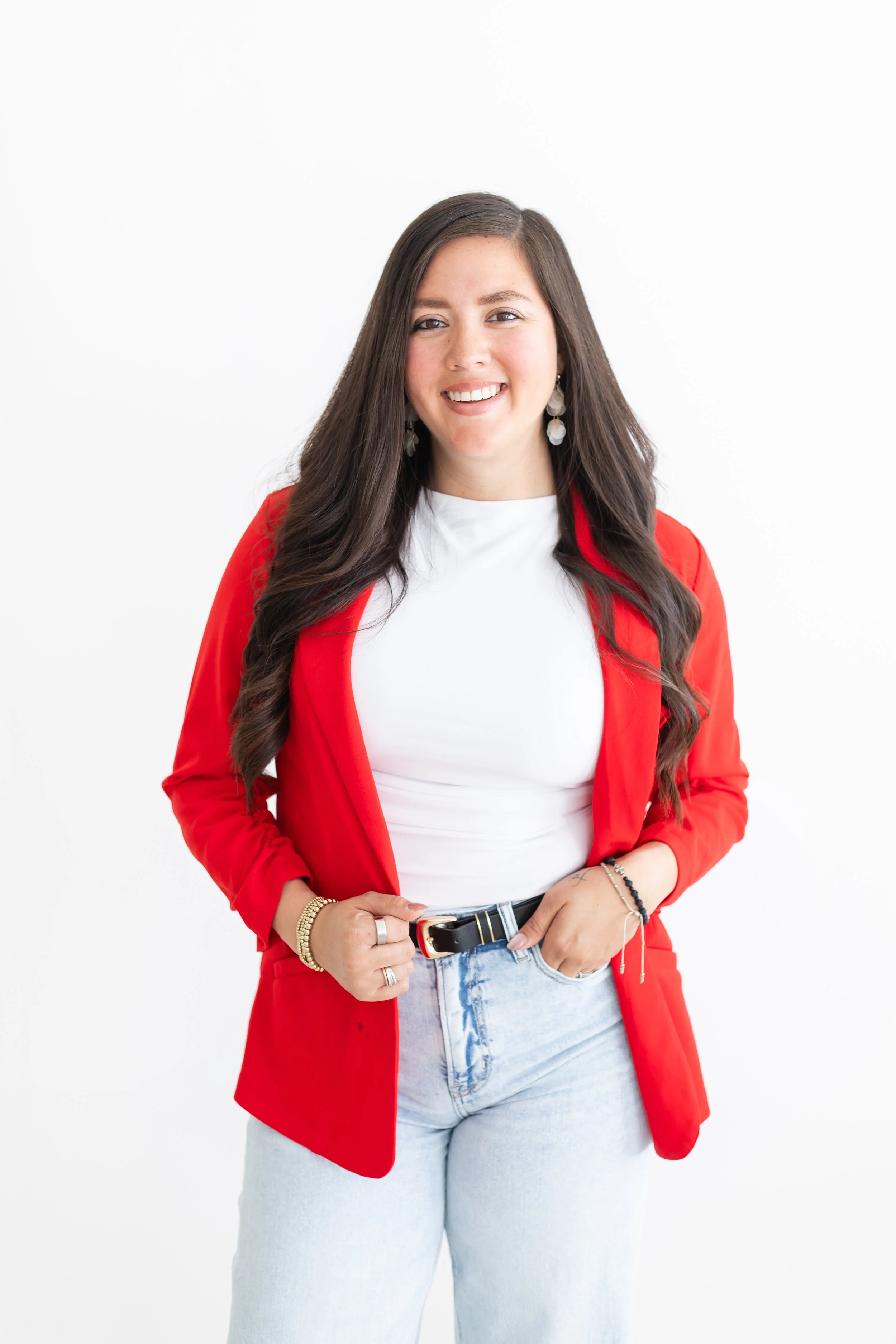 A woman with long dark hair wearing a red jacket, white shirt, and light blue jeans, smiling and posing against a white background.