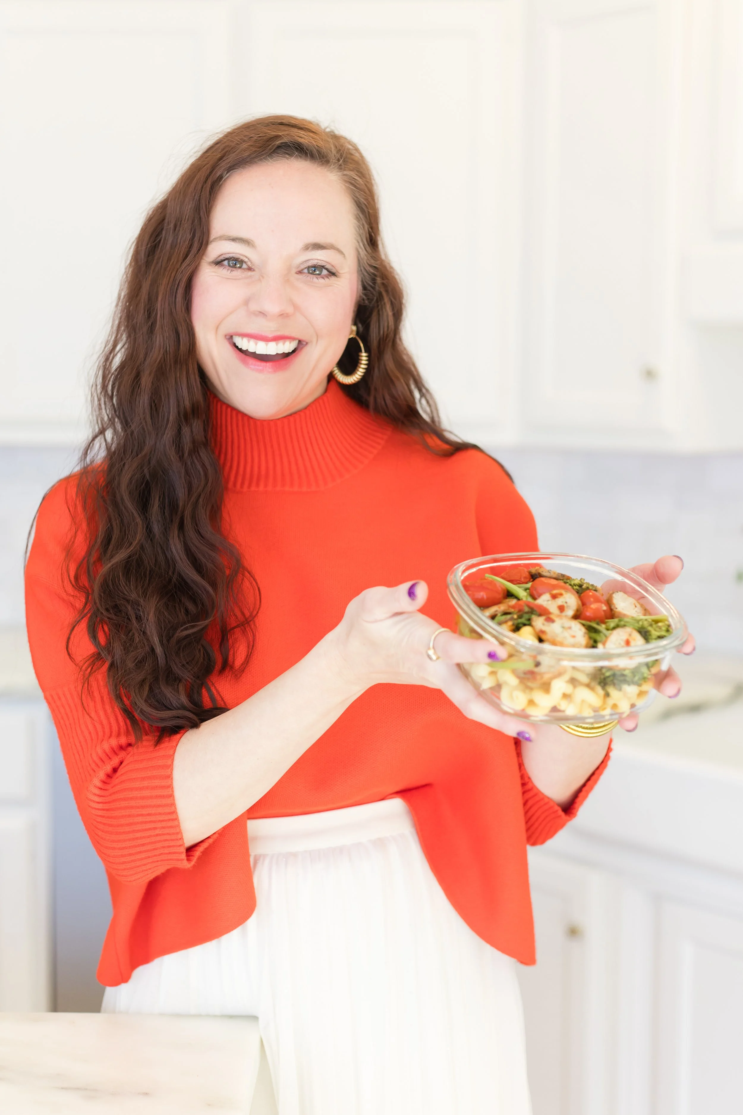 A woman with long wavy brown hair smiling and holding a clear glass bowl of salad in a kitchen.