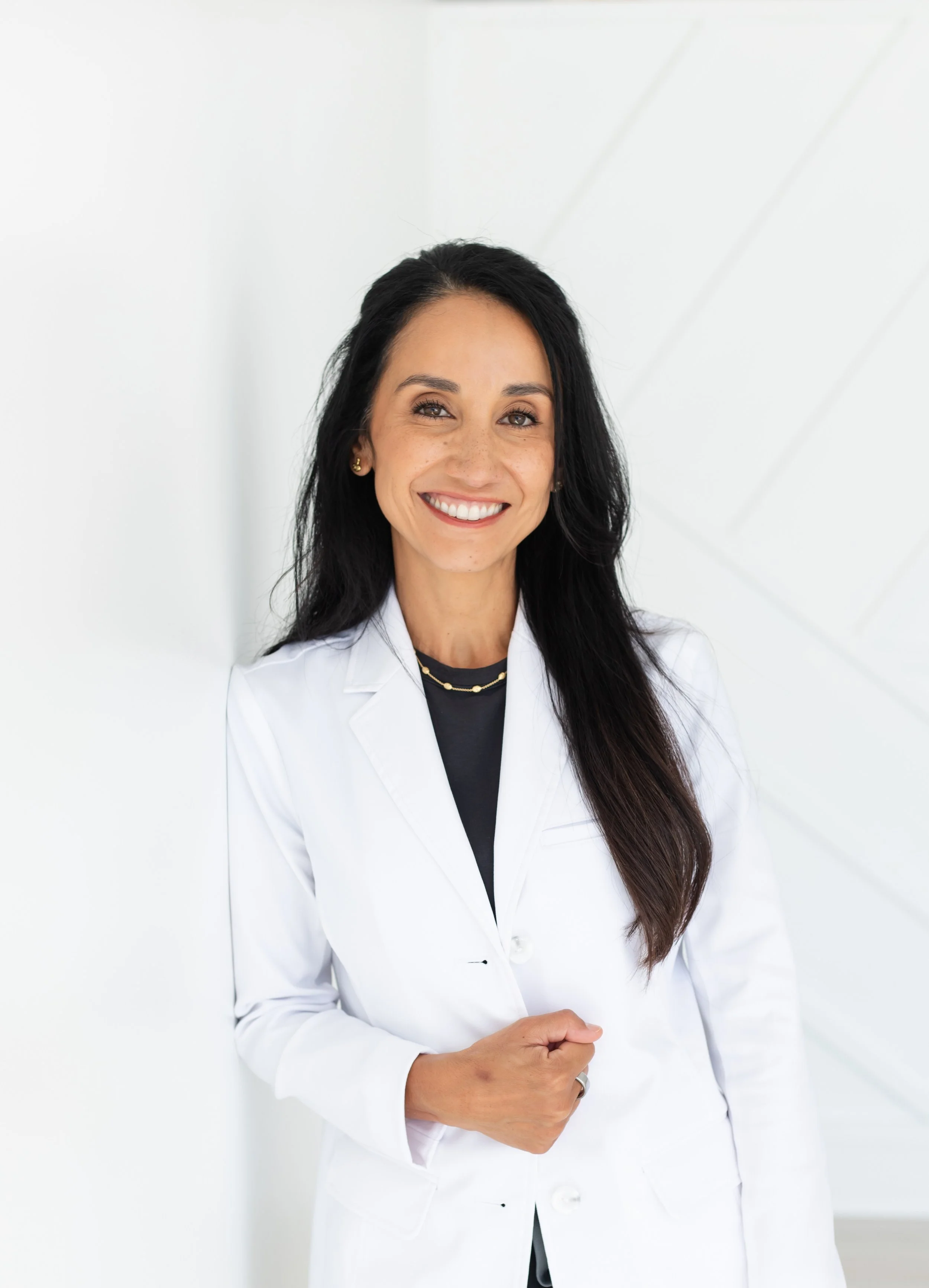 A woman with long dark hair and a big smile, wearing a white lab coat, standing against a white wall.