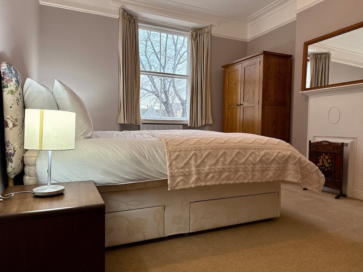 A cozy bedroom with a bed, pillows, and a cream-colored blanket. There is a wooden nightstand with a lamp, a window with curtains showing trees outside, a wooden wardrobe, and a large mirror on the wall.