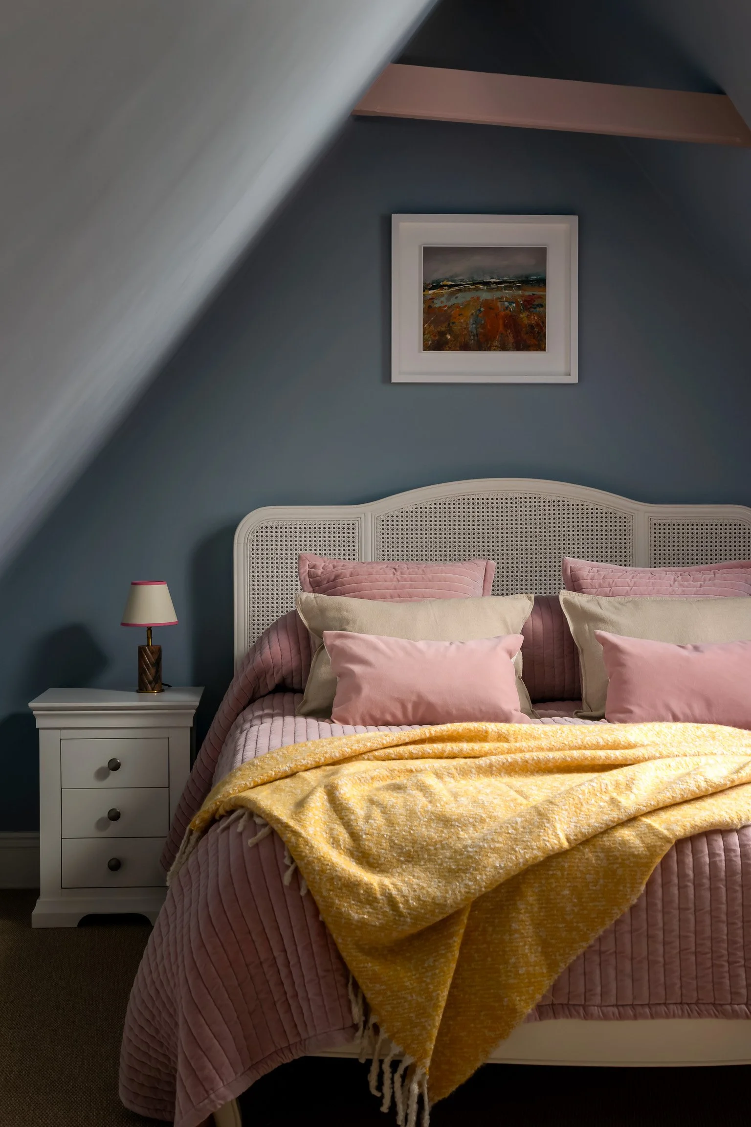 A cozy bedroom with a bed featuring pink, beige, and cream pillows, a pink quilt, and a yellow throw blanket. There is a white nightstand with a small lamp, and a framed landscape painting hangs on a blue wall above the bed. The room has a sloped cei