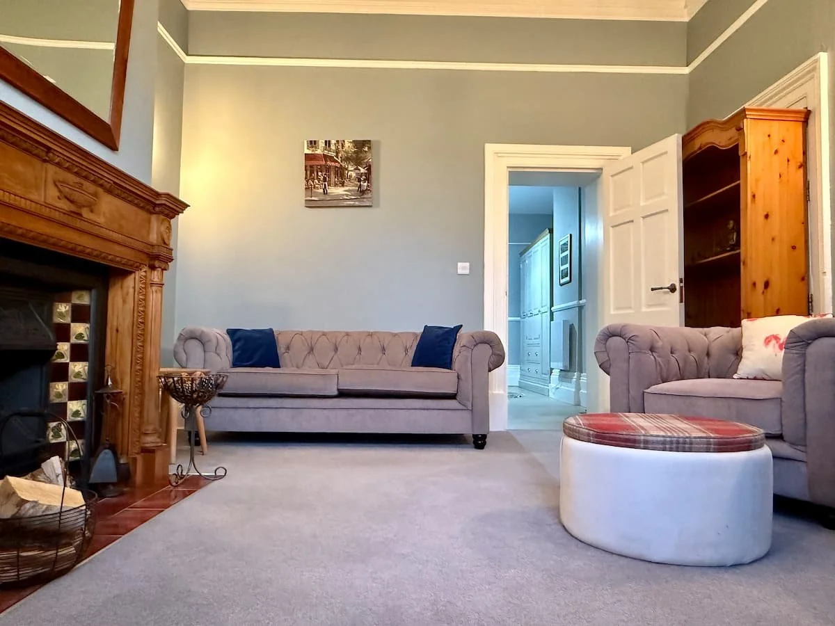 Living room with blue walls, a fireplace with wooden mantel, two gray sofas with tufted arms, a plaid ottoman, and a doorway leading to a hallway.