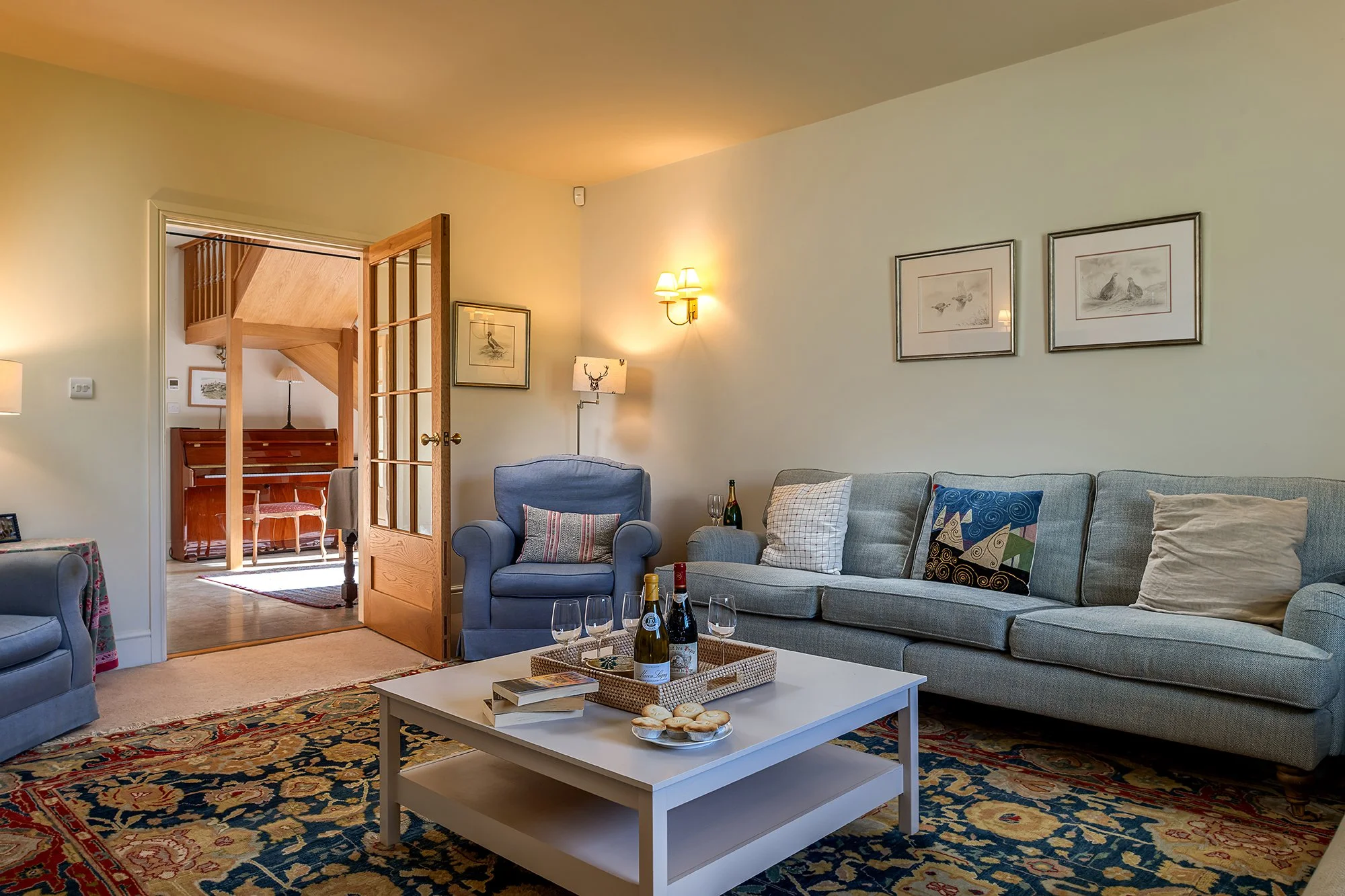 Living room with blue armchair, gray sofa with decorative pillows, coffee table with wine bottles and snacks, framed wall art, wooden door open to a room with a piano, and a patterned rug.