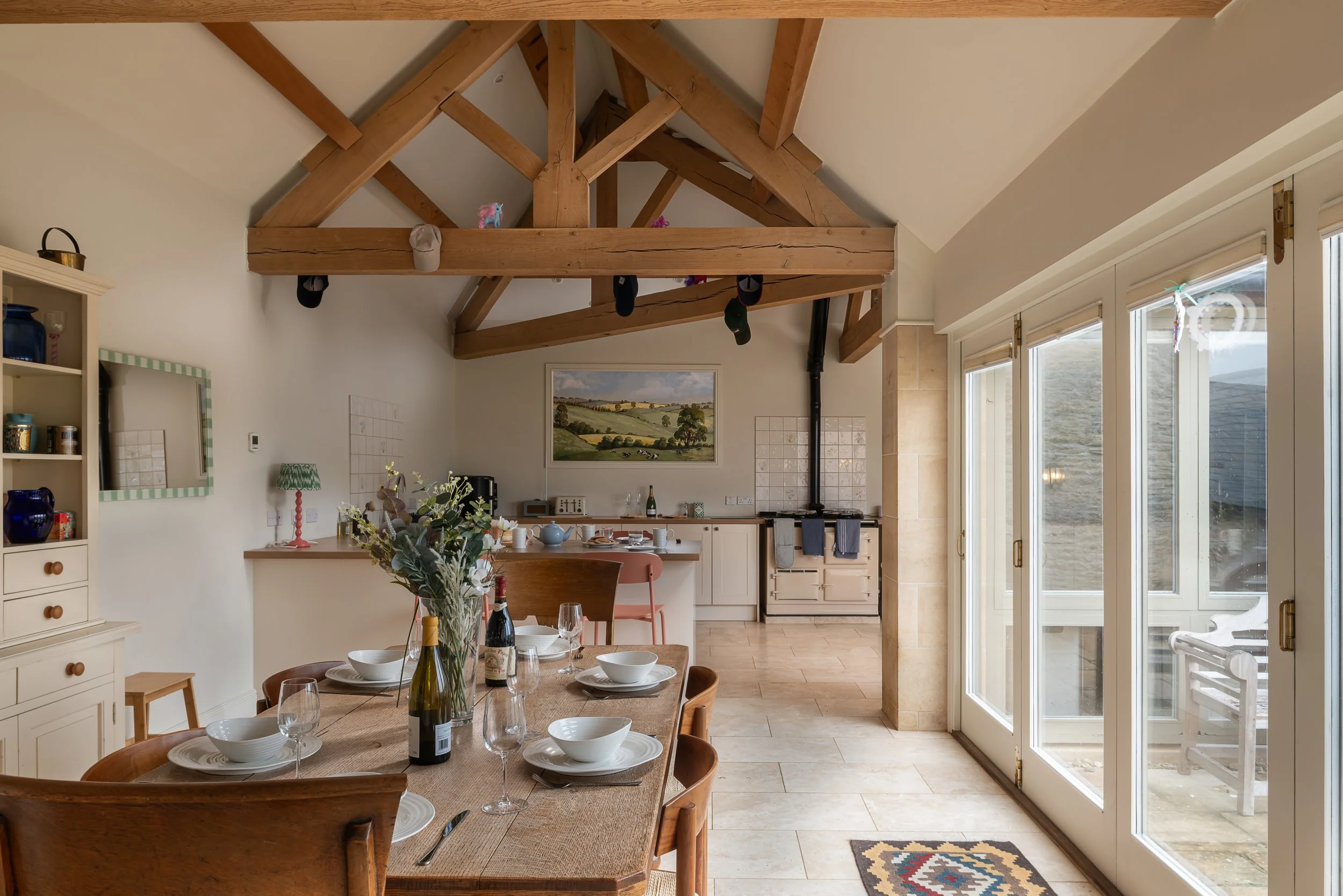 Dining area with wooden table set for four, wine bottles, and glasses, in front of kitchen with cream cabinets, stove, and artwork. Large glass doors leading outside, exposed wooden beams on ceiling.