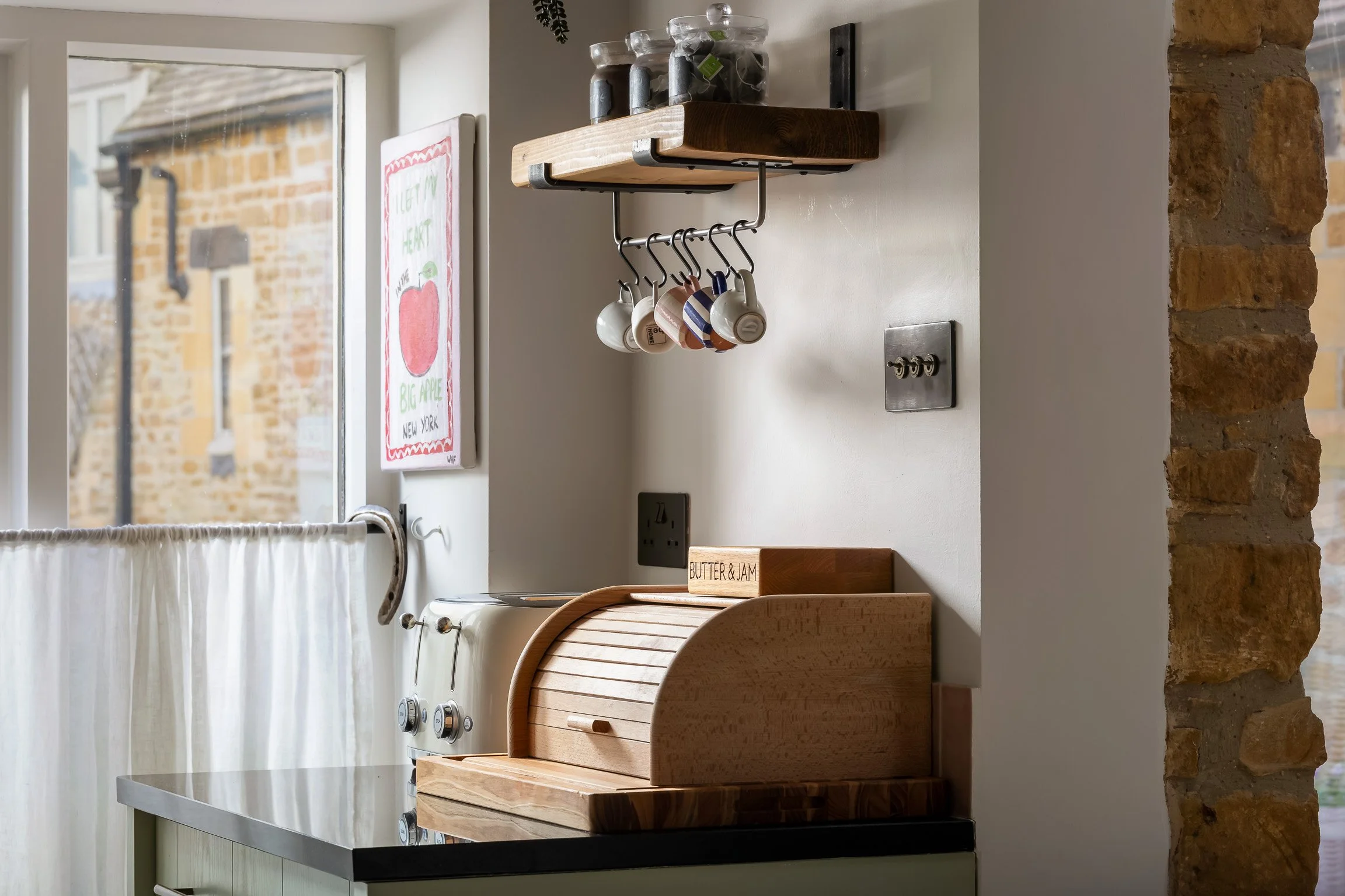 Kitchen corner with wooden bread box, wall-mounted shelf holding jars, mug hooks with mugs, nearby window with cream-colored curtain, and brick wall on the right.