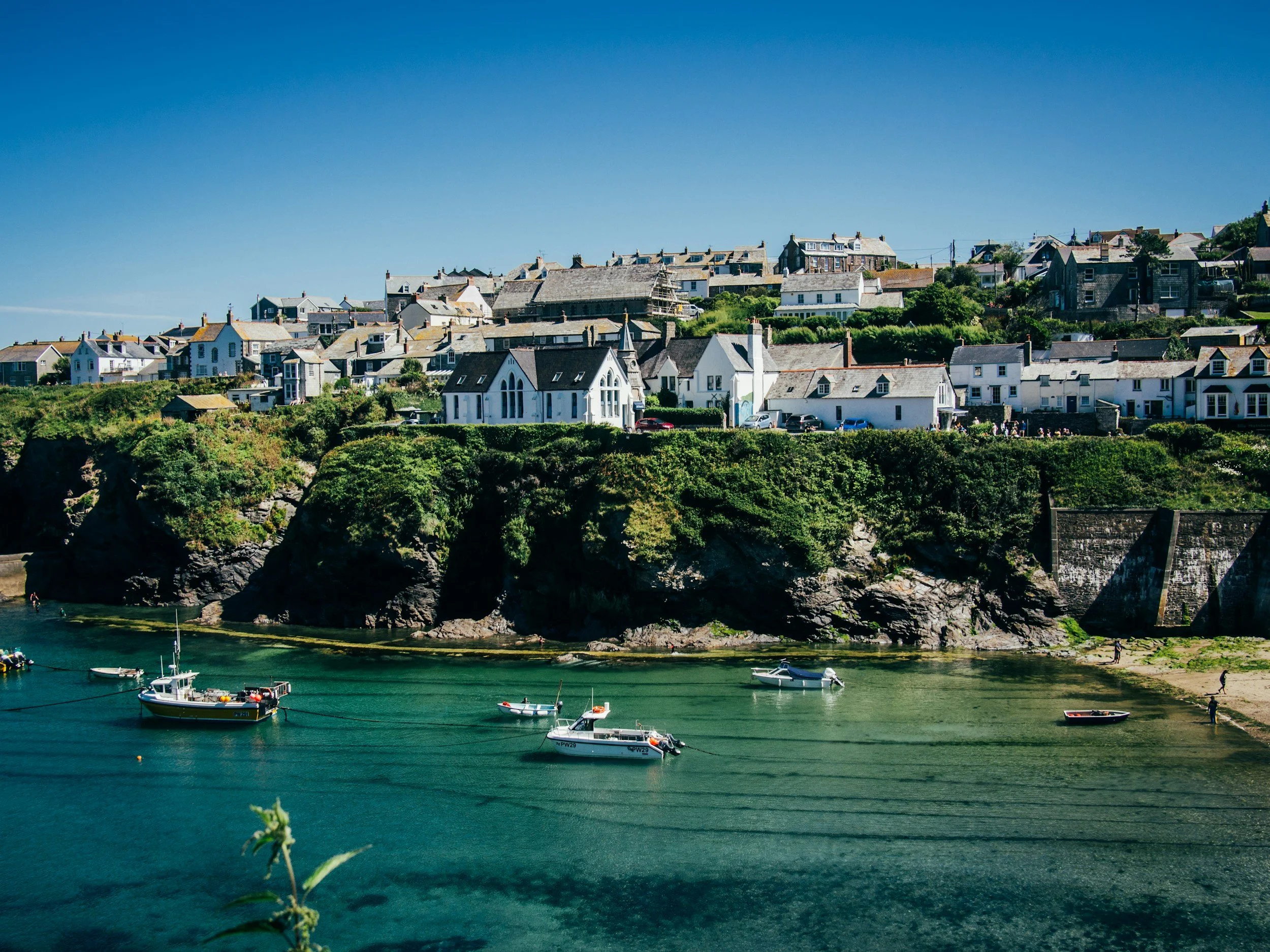 A coastal scene with boats floating in clear, turquoise water, and a hillside dotted with white and gray houses and green foliage against a bright blue sky.