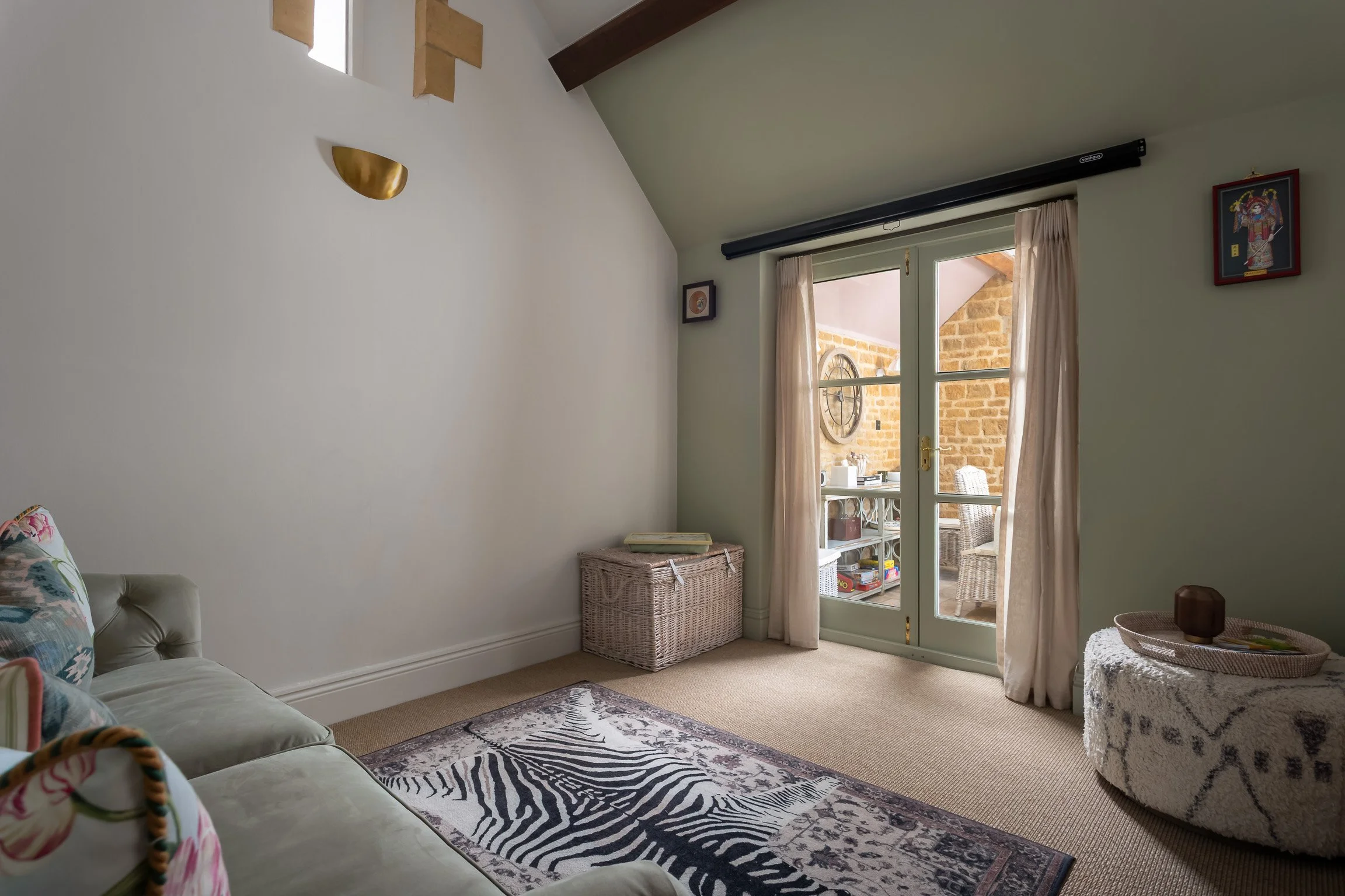 Cozy living room with a sofa and colorful cushions, a patterned rug, a wicker storage chest, and a glass door leading to a balcony with outdoor furniture and a brick wall.