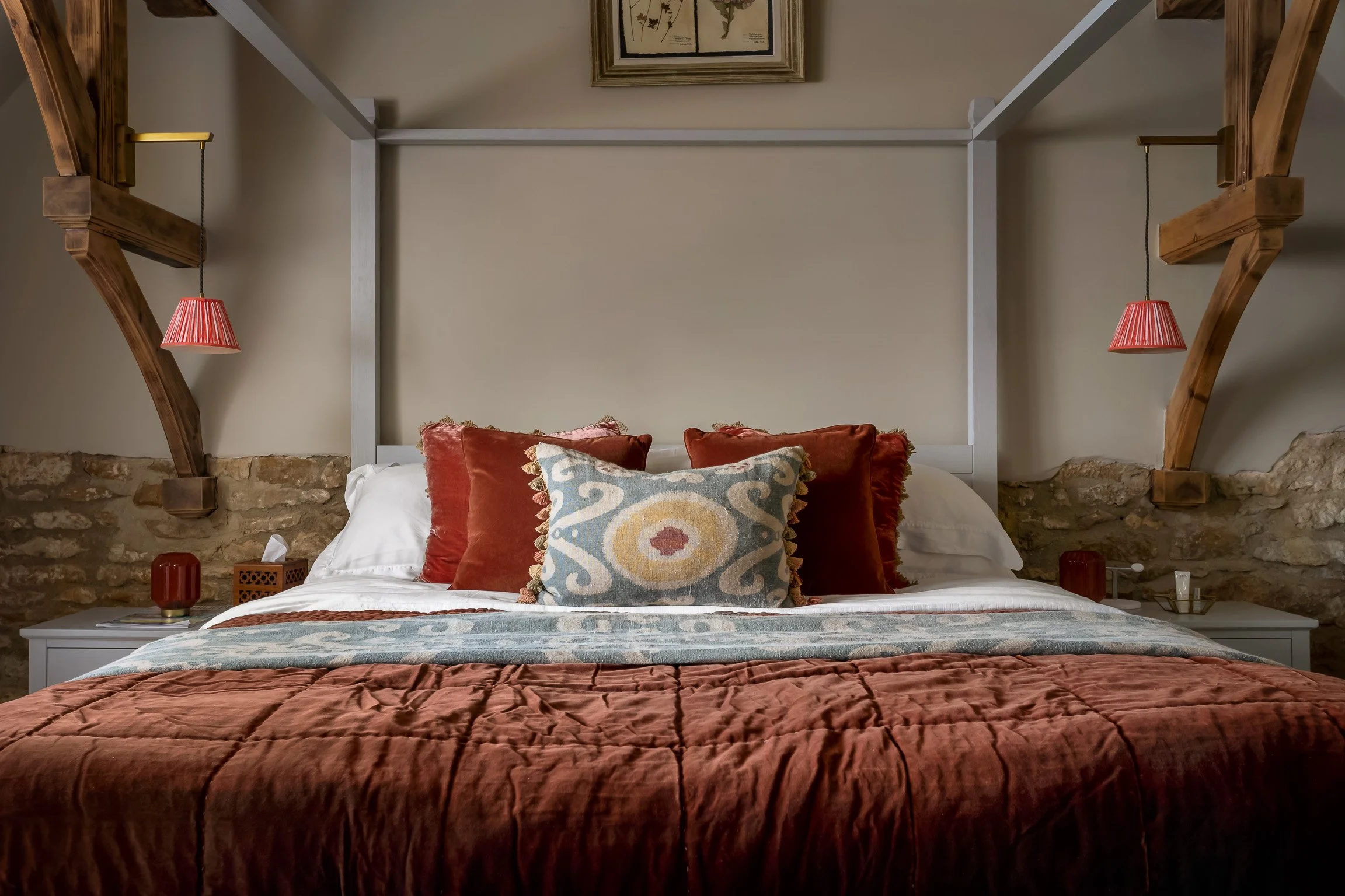 A neatly made bed with a rust-colored quilt, decorative pillows, and a patterned runner. Two bedside tables with lamps are on either side, and a framed picture hangs above the bed. The room features exposed stone walls and wooden beams.