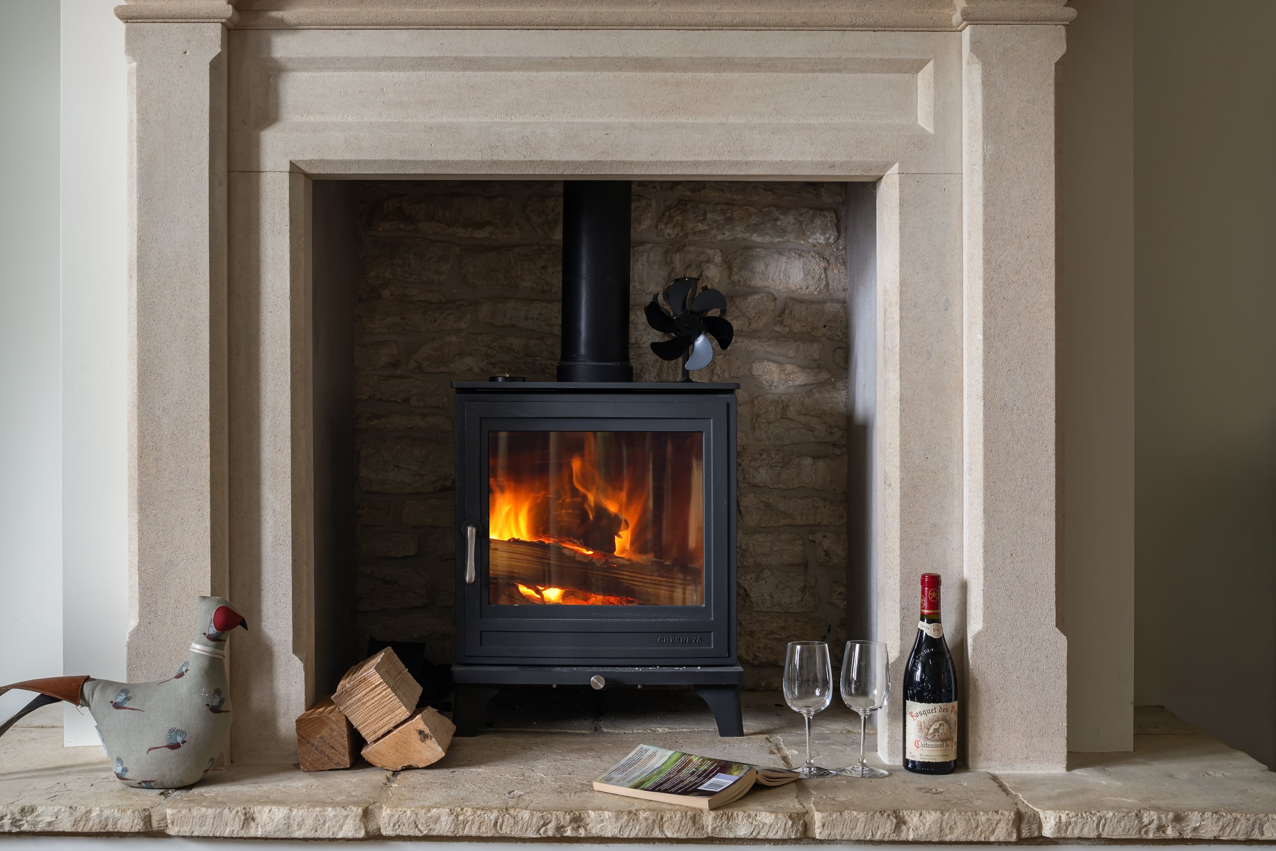 A cozy fireplace with a fire burning, decorated with a fabric duck, firewood, a bottle of wine, two empty wine glasses, and a book on the stone hearth.
