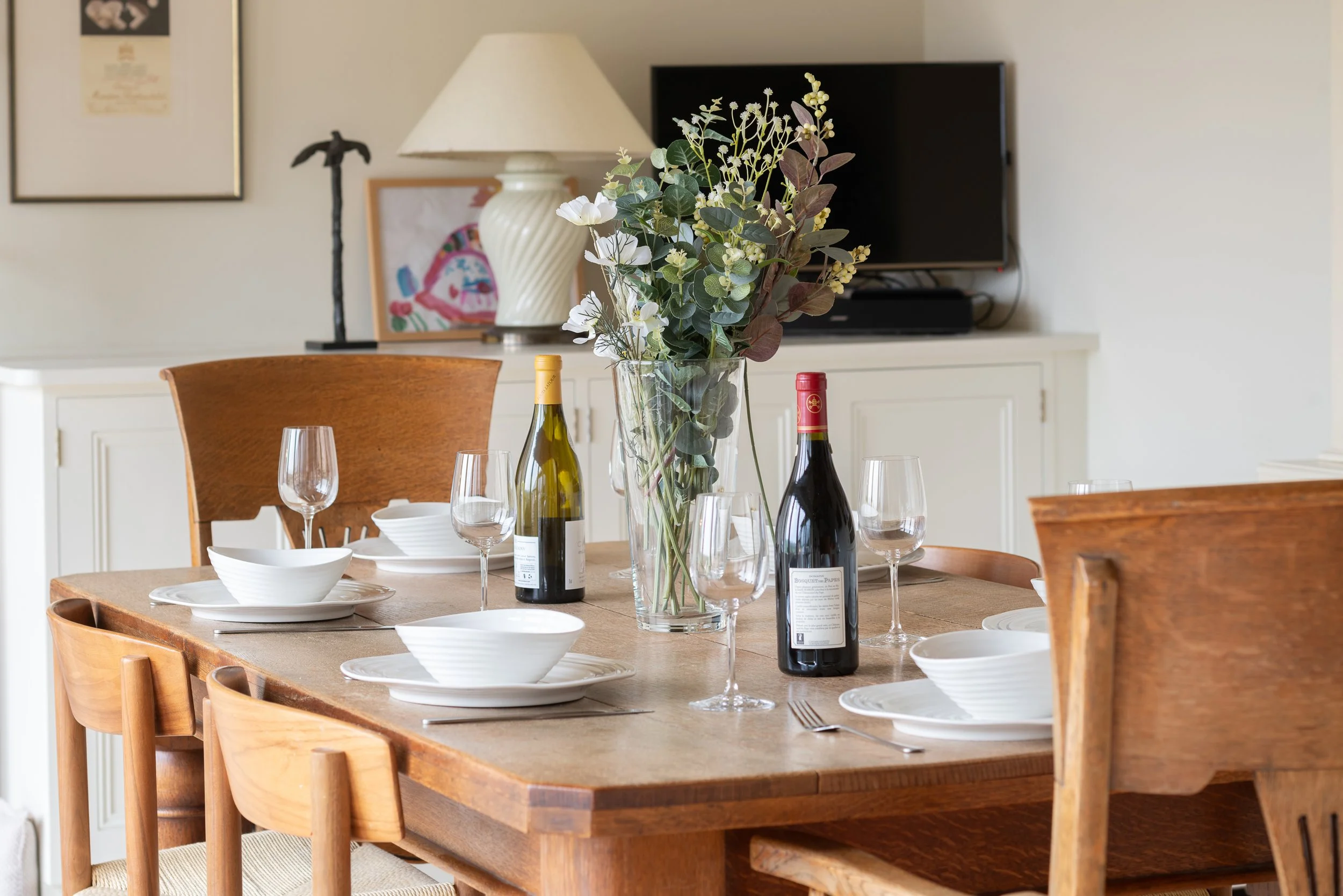 A dining room table set with white bowls, plates, wine glasses, and two bottles of wine. There is a large vase with green and white flowers in the center of the table. In the background, a sideboard with framed artwork, a lamp, a small sculpture, and