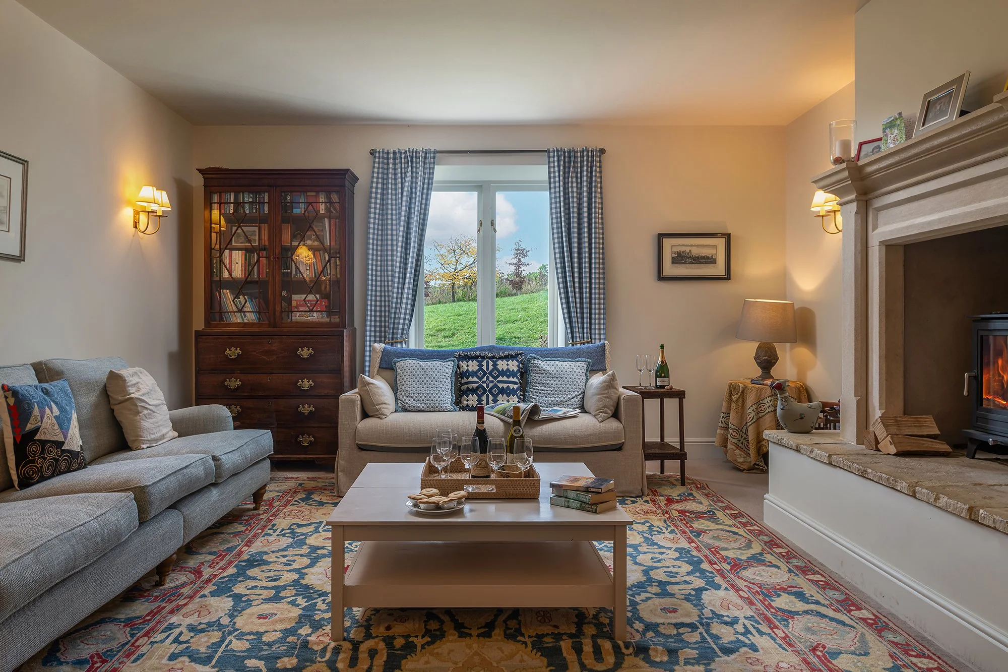 A cozy living room with two sofas, a coffee table, a fireplace, a wooden bookcase, a window with checkered curtains, lamps, and decorative items.