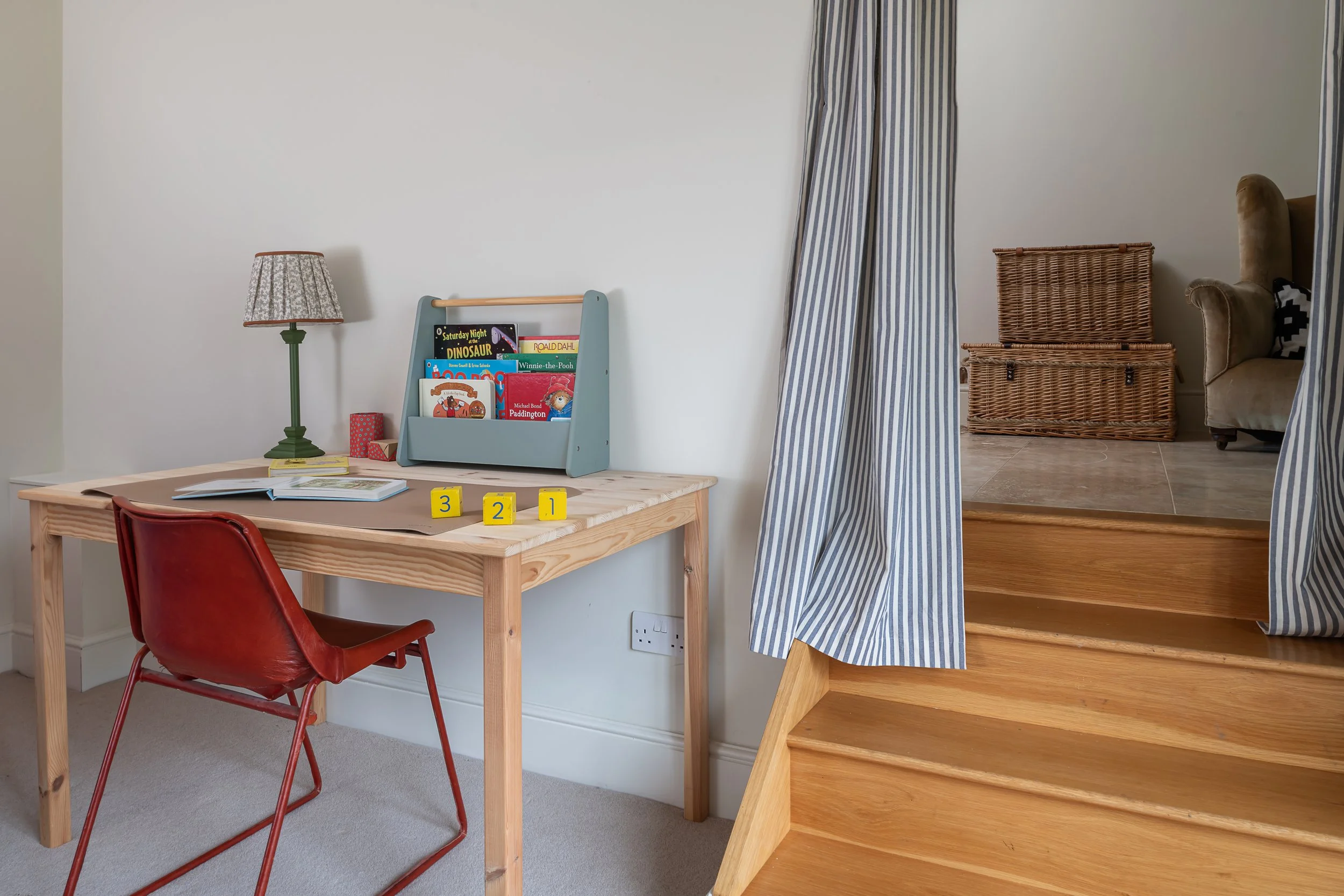 Child's playroom with a wooden desk, red chair, bookshelf with children's books, table lamp, and wooden steps with blue-striped curtains leading to an upstairs area with a wicker basket and sofa.