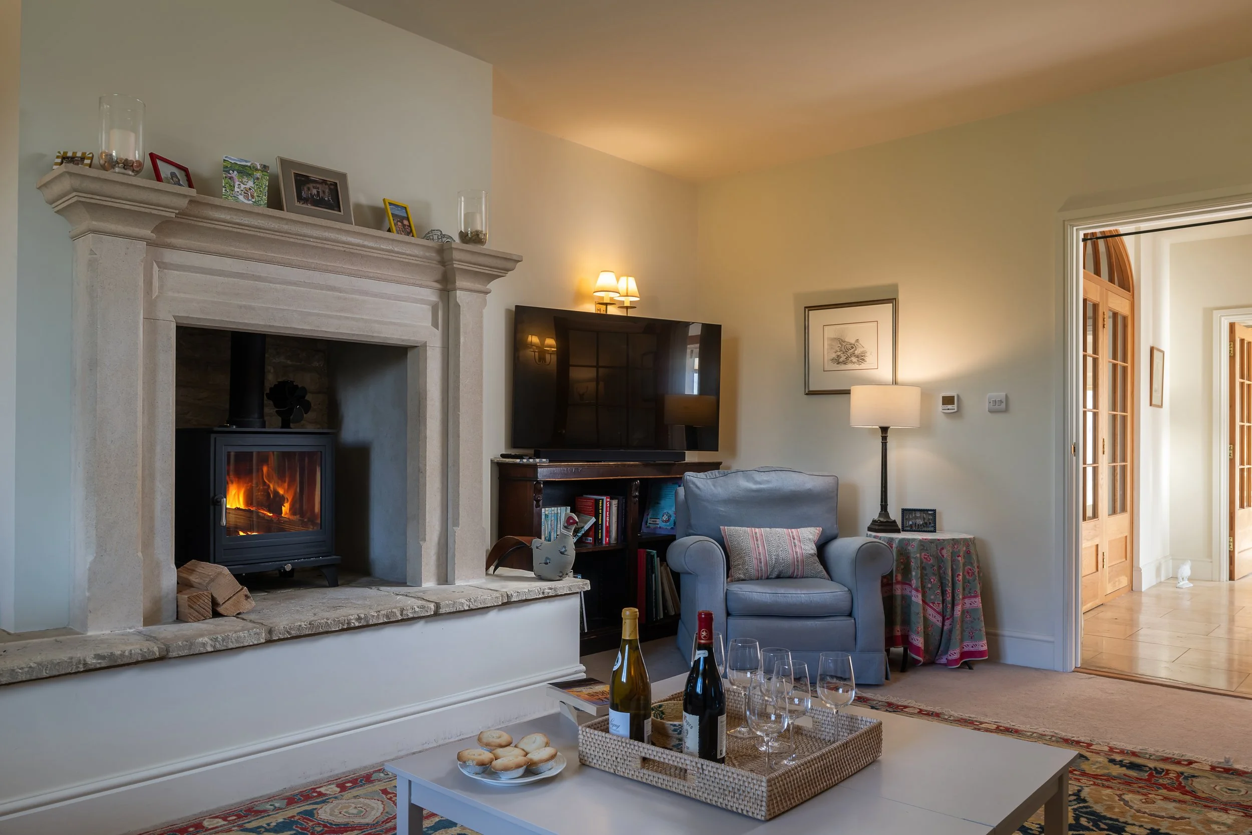 Living room with a lit fireplace, a gray armchair with striped pillow, a flat-screen TV on a wooden stand, a side table with a lamp and framed picture, and a table with wine bottles and glasses.