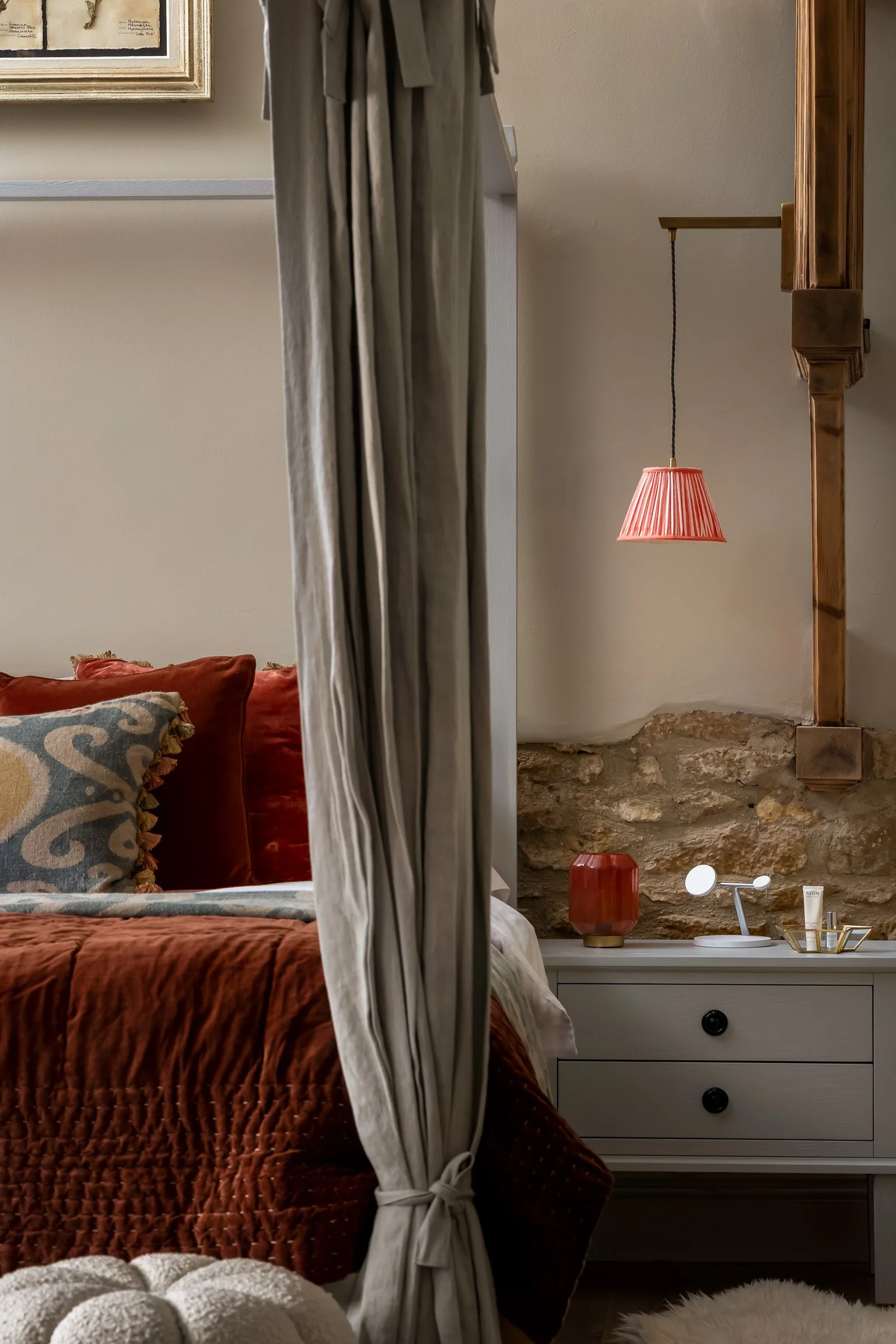 A cozy bedroom with a bed, rust-colored quilt, decorative pillows, and a close-up of a bedside table with a red lamp, mirror, and skincare products. The room has a stone wall and a hanging pink lampshade.