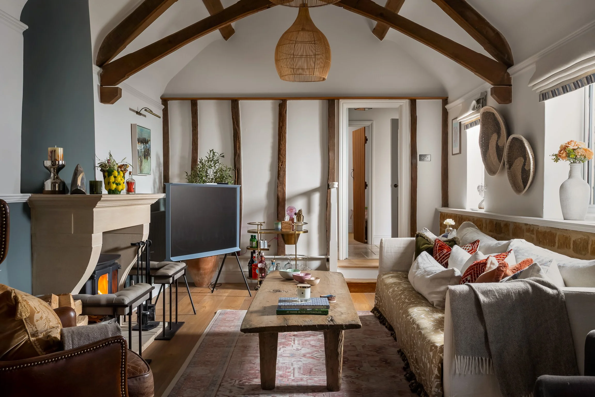 Cozy living room interior with exposed wooden beams, a stone fireplace, a large cream-colored sofa with assorted pillows, a rustic wooden coffee table, a TV, decorative baskets and vases, ceiling pendant light, and windows with striped curtains.