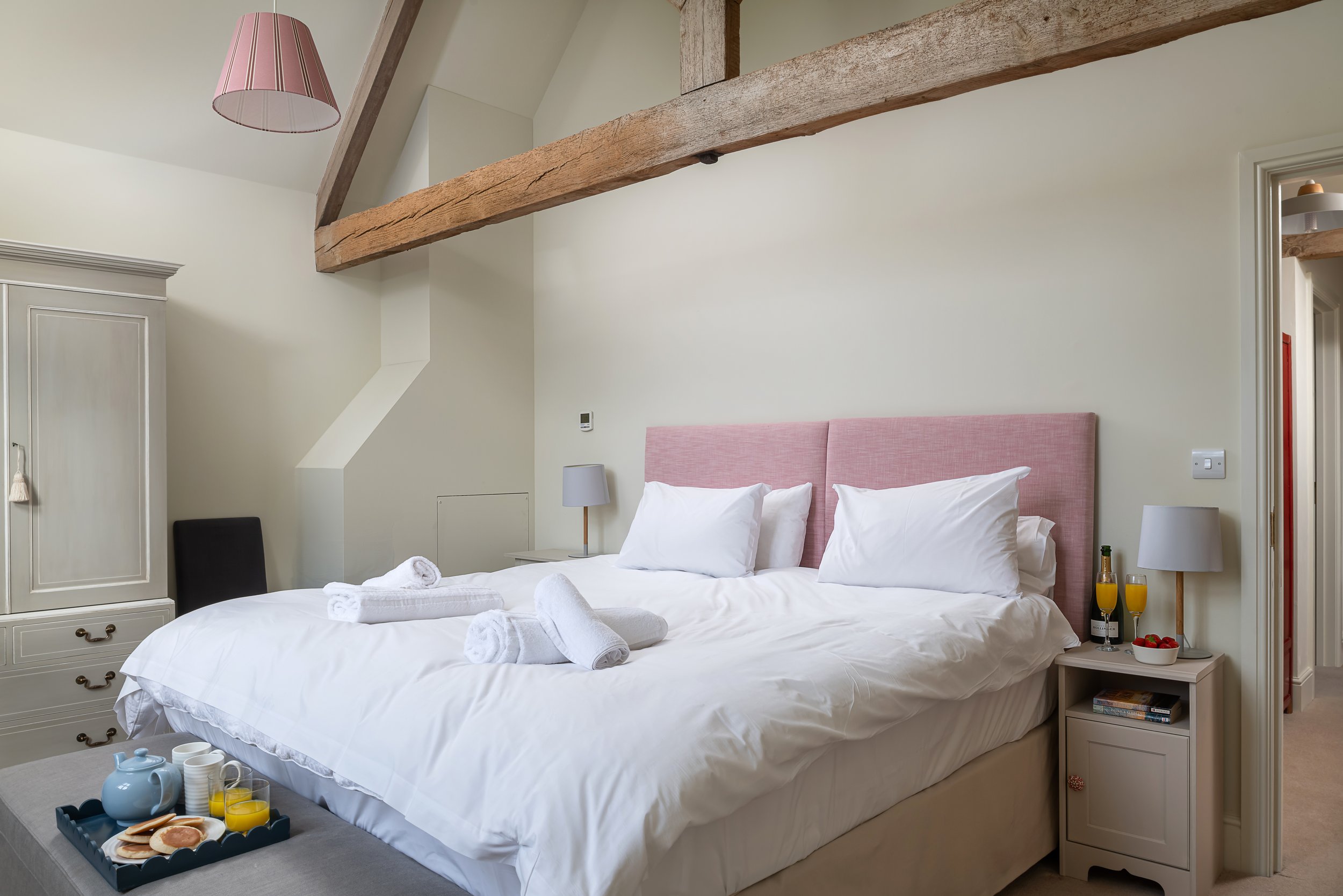 A cozy hotel bedroom with a large bed, pink headboard, white bedding, rolled towels, bedside tables with lamps, and breakfast tray.