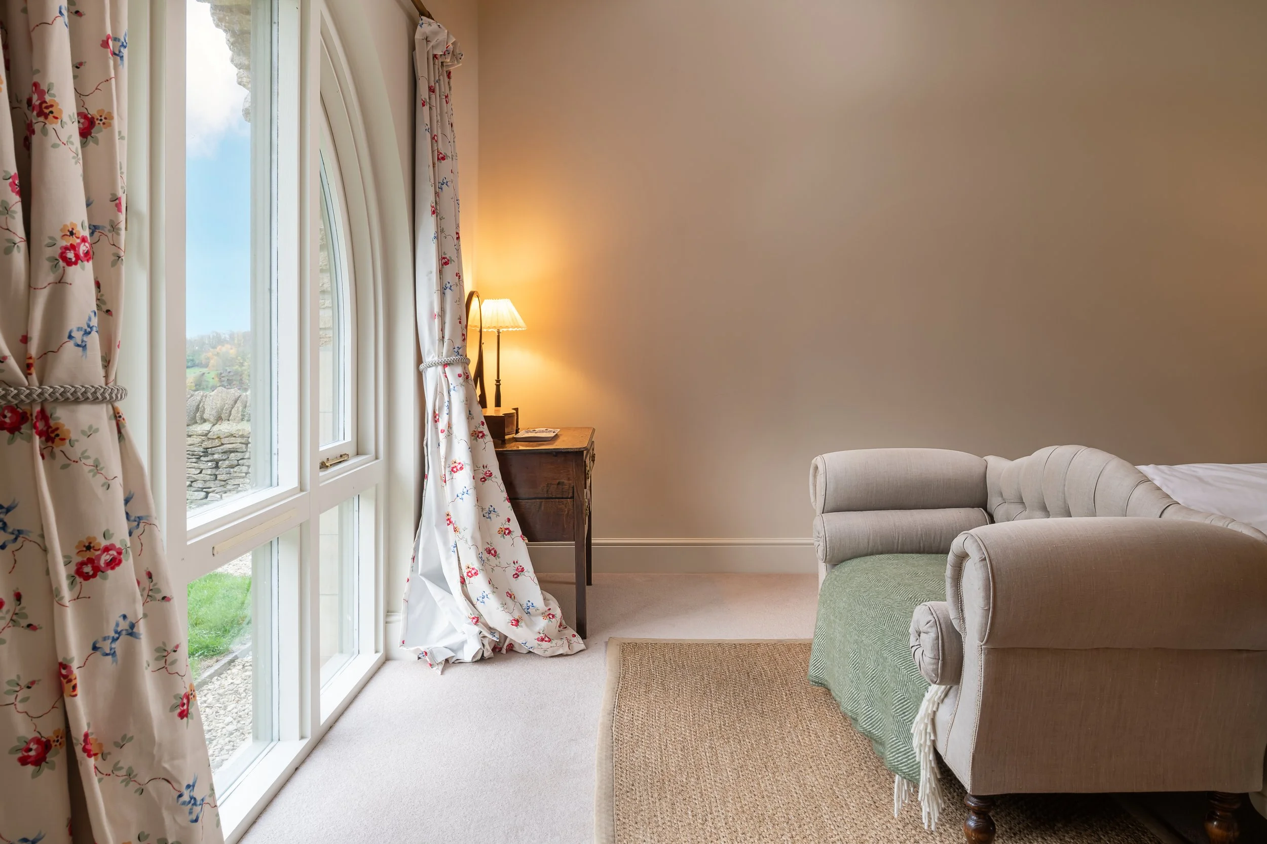 A cozy bedroom corner with a large glass window covered by floral curtains, a small wooden nightstand with a lamp, and a white curved sofa with a green blanket.