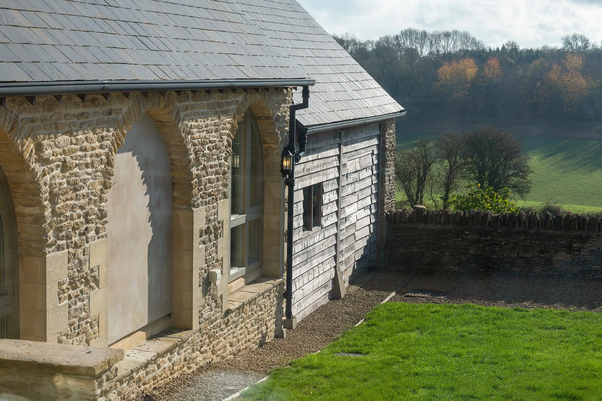 Side view of a stone house with arched windows and a wooden extension, overlooking a lush green field and trees in the distance.