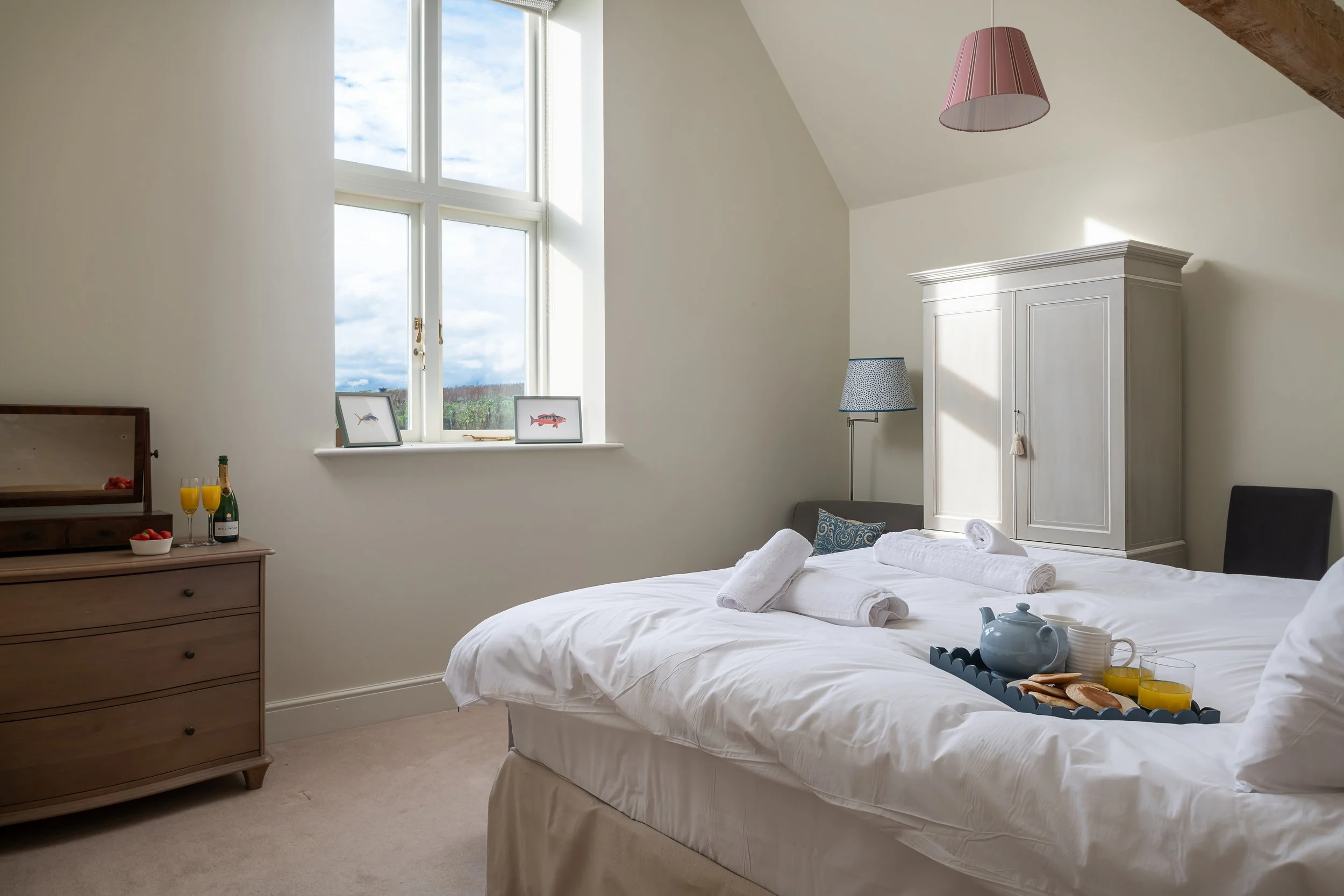 Bright bedroom with large window, white bedding, towels, breakfast tray, wardrobe, and armchair.