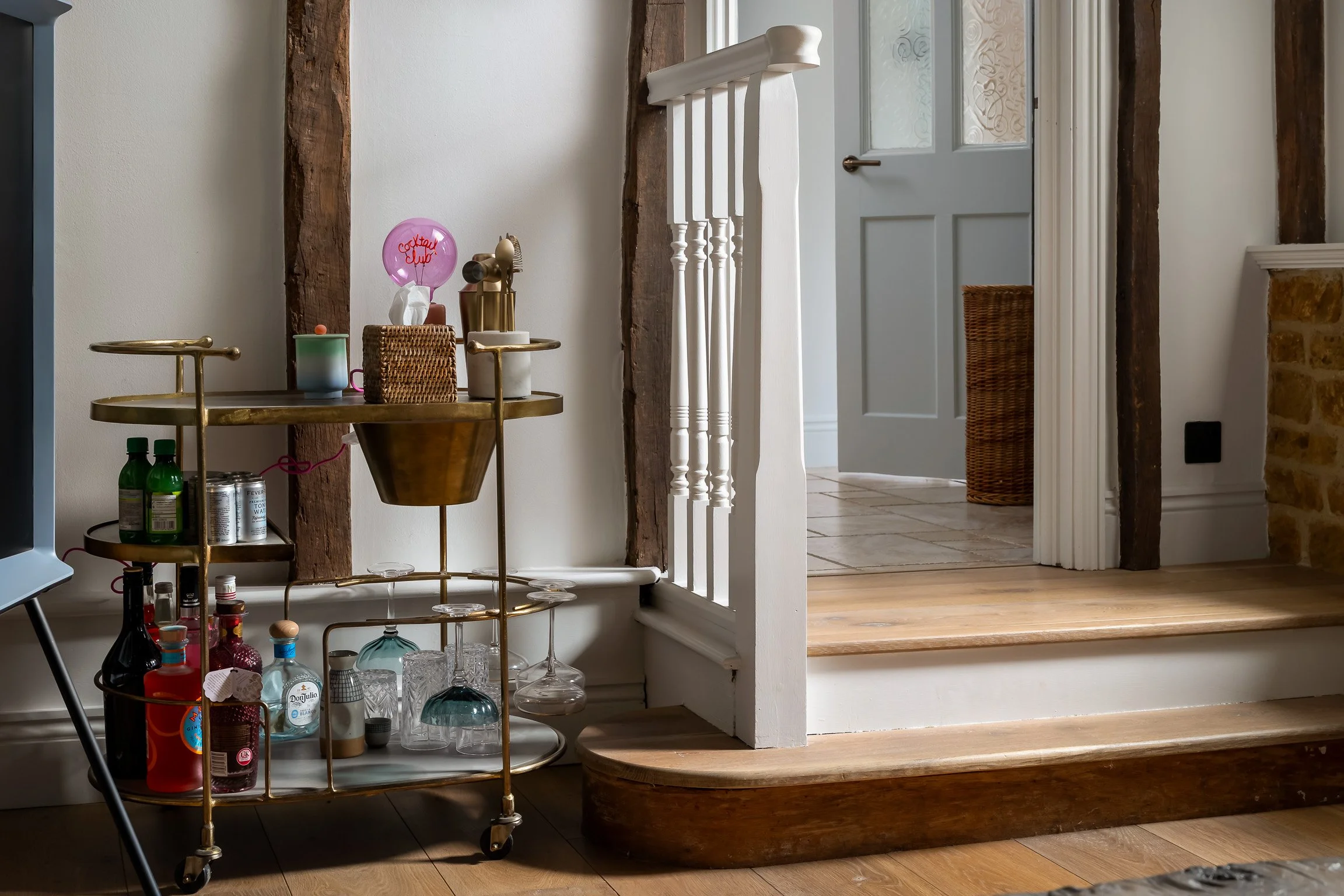 A brass bar cart with bottles, glasses, and decor, positioned next to a white wall with a rustic wooden beam, and a doorway leading to a tiled room with a woven basket. The room has wooden floors and trim.