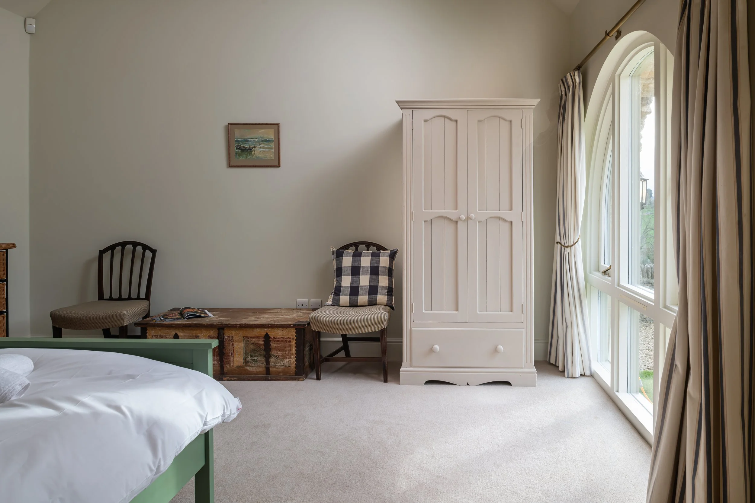 A bright bedroom corner with a bed, two chairs, a wooden trunk, a white wardrobe, and large windows with curtains.
