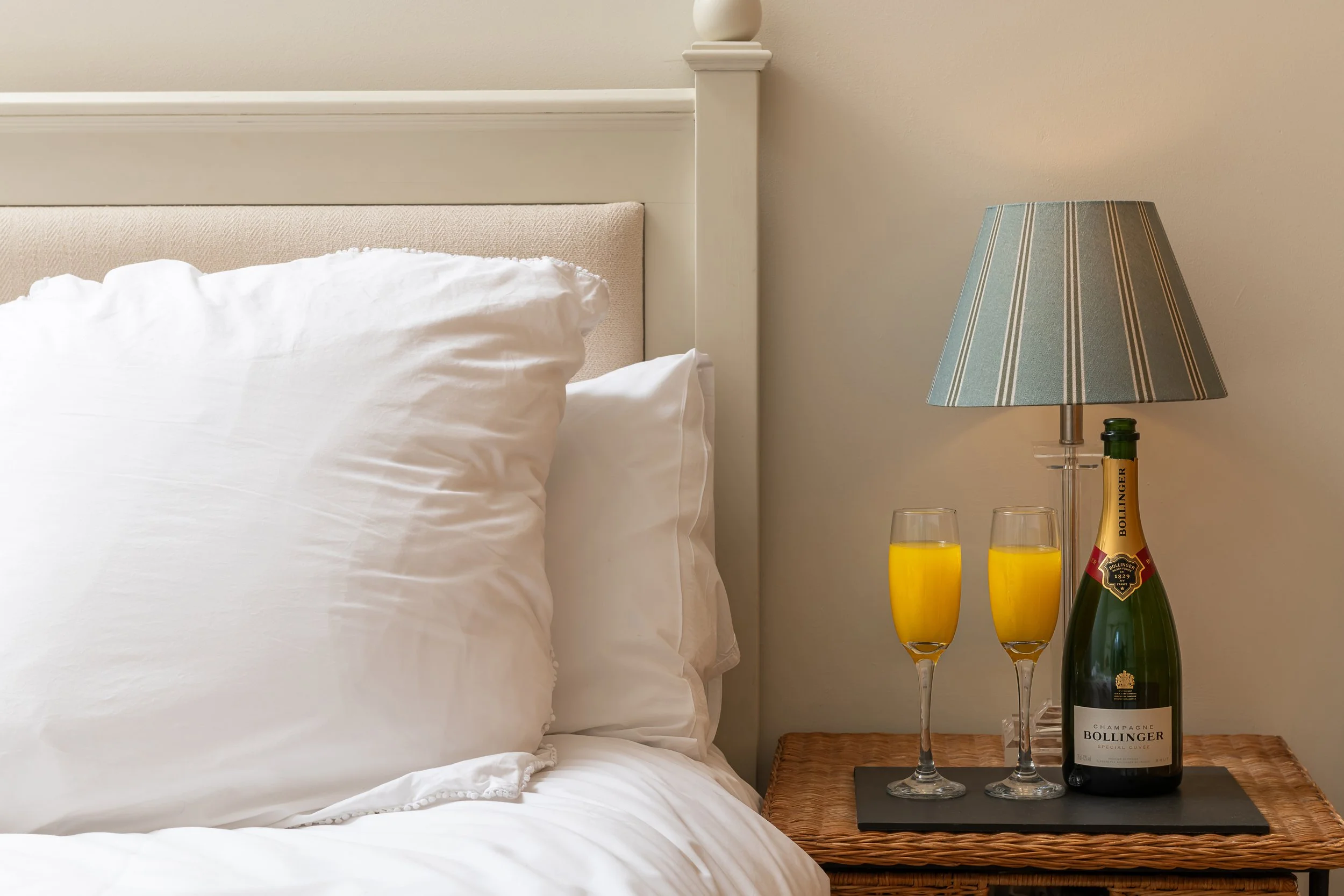 A bedroom nightstand with a bottle of Bollinger champagne, two glasses filled with orange juice, and a patterned lamp, beside a bed with white pillows and bedding.