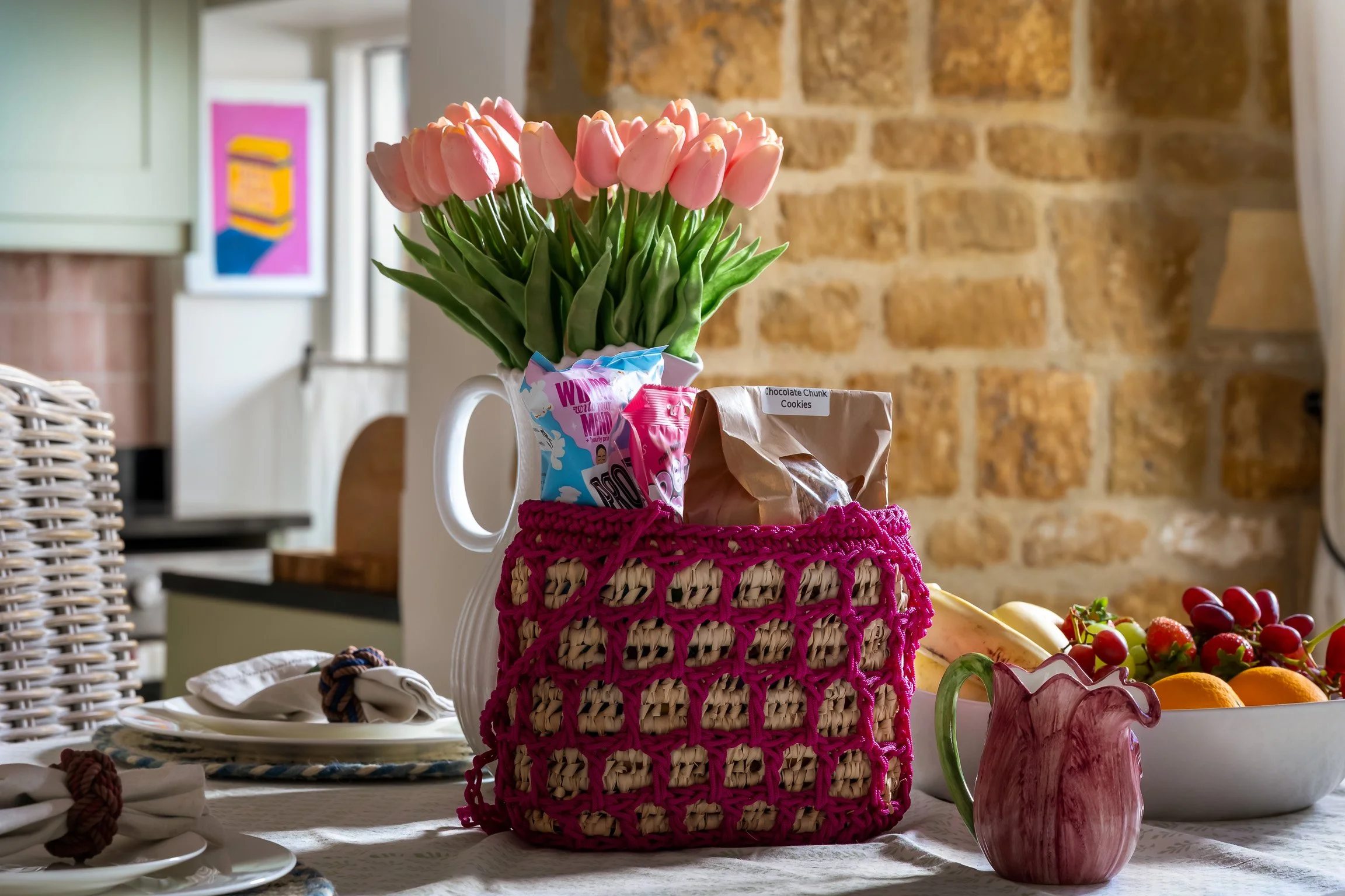 A table decorated with a vase of pink tulips, a pink woven bag holding snacks, a fruit bowl with bananas, grapes, and oranges, and a pink pitcher, with a brick wall and window in the background.