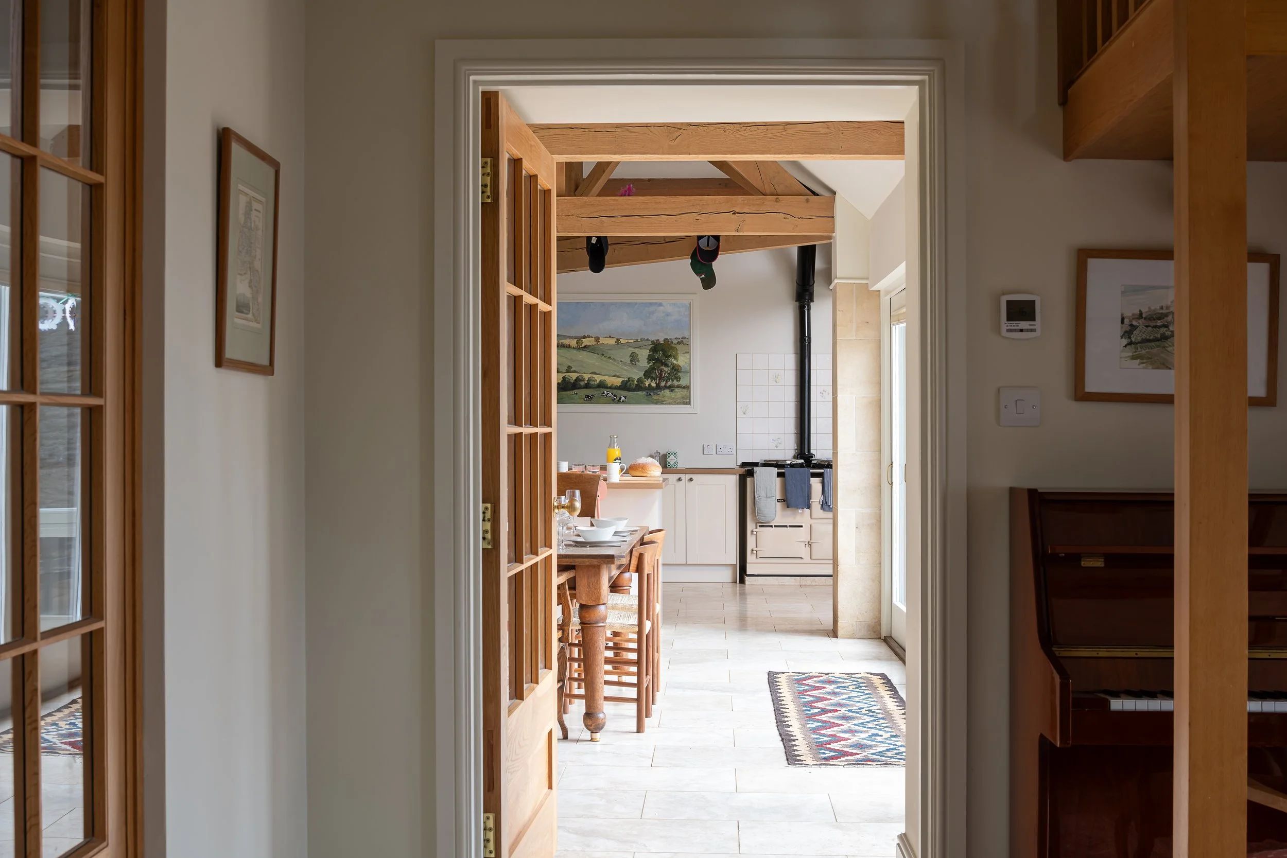 View through doorway into a kitchen with a wooden dining table, chairs, a painting, and a stove with a black chimney pipe.