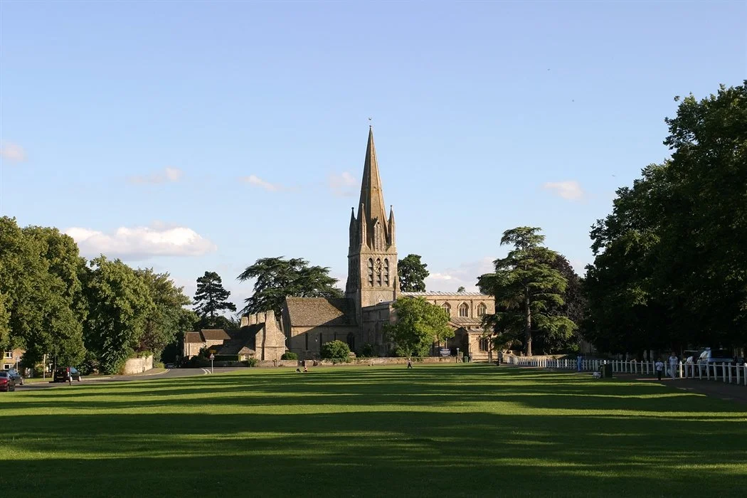 A historic stone church with a tall steeple surrounded by a grassy park with trees and a few people walking.