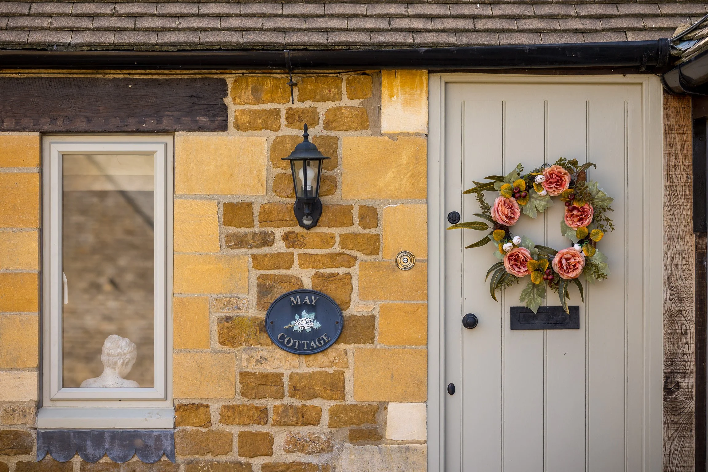House entrance with a beige door decorated with a floral wreath, a window to the left with a white frame and sculpture inside, and a black lantern mounted on a brick wall that says 'May Cottage'