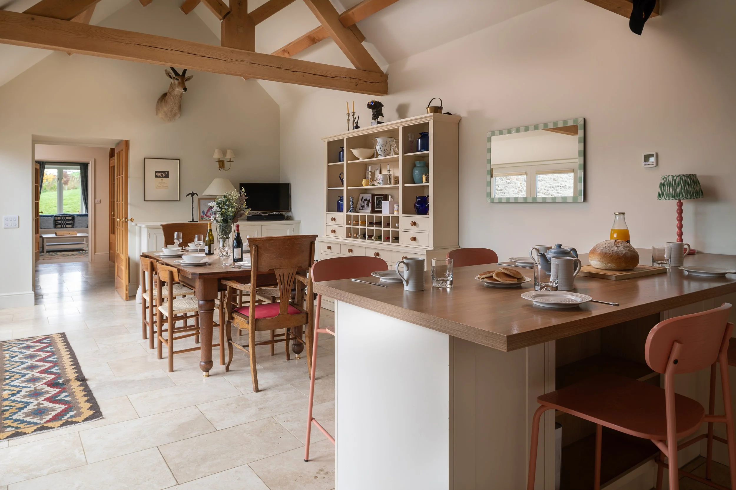 A cozy dining area with a wooden table set for breakfast, surrounded by chairs, and a kitchen island with a pink chair. A wooden shelving unit on the wall holds plates, bowls, and decorative items. A mounted animal head is on the wall, and a doorway 