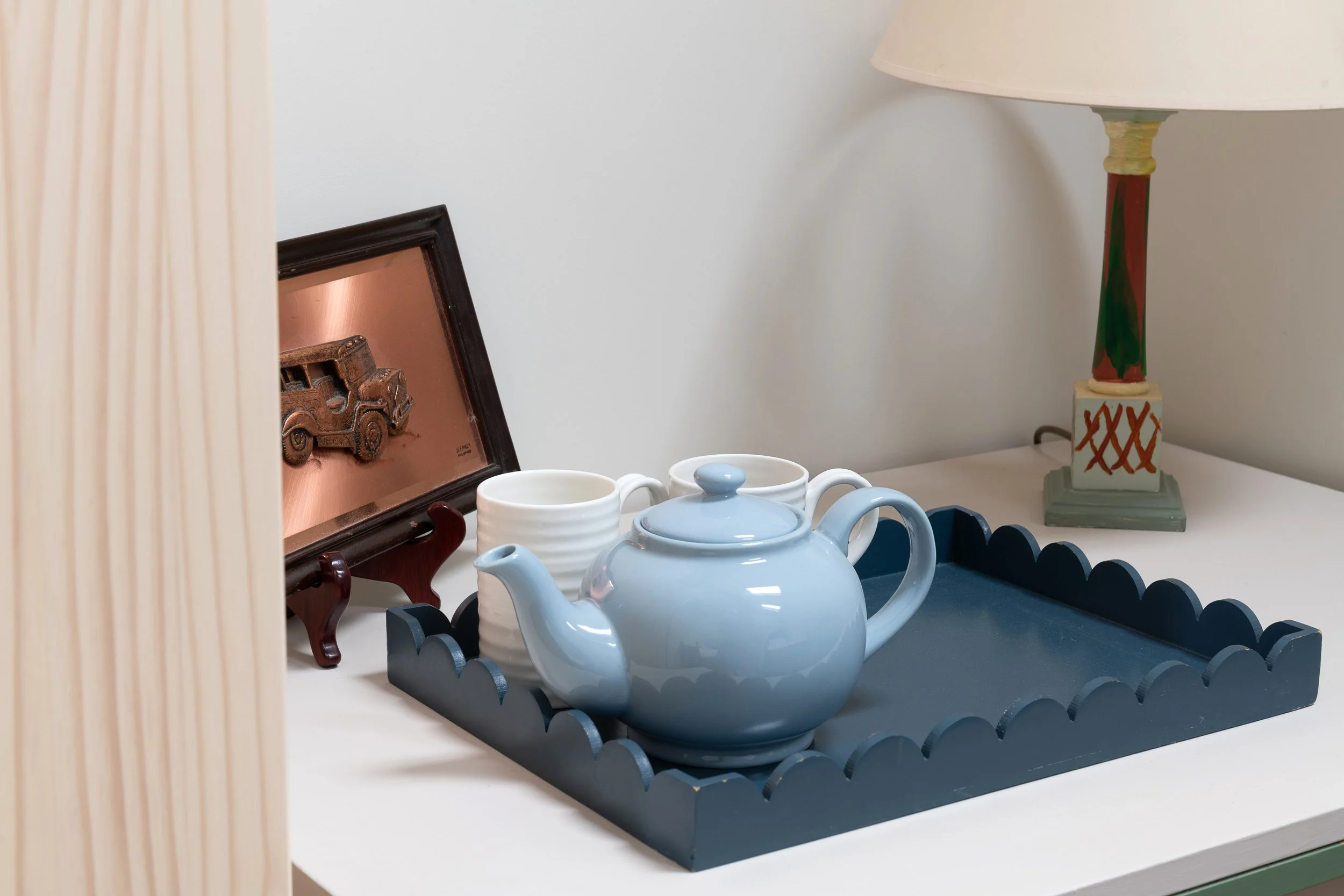 Tea pot with cups on a tray, picture frame with car, and lamp on a white table.