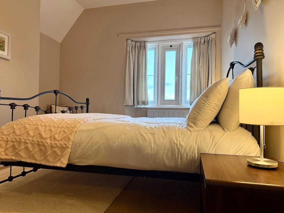 A cozy bedroom with a metal bed frame, white bedding, pillows, a wooden nightstand with a lamp, and a window with curtains showing a cloudy sky.
