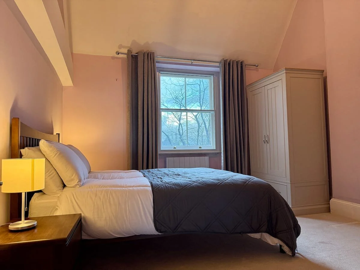 A cozy bedroom with a bed, nightstand, lamp, window with curtains, and a wardrobe.