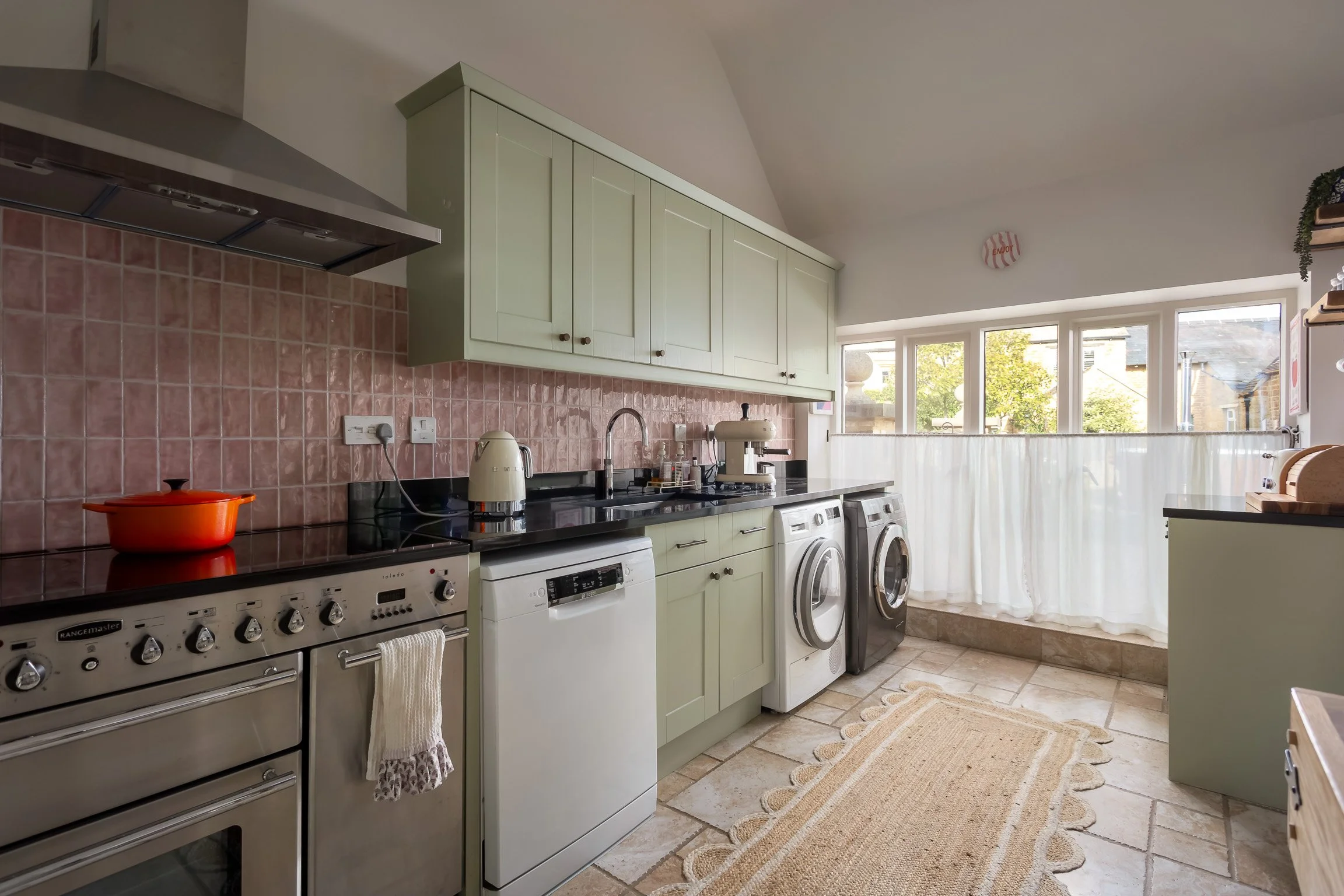 A kitchen with green cabinets, pink tiled backsplash, black countertop, and window with white curtains. Appliances include a stove, dishwasher, washing machine, and coffee maker. There is a beige rug on the tiled floor.