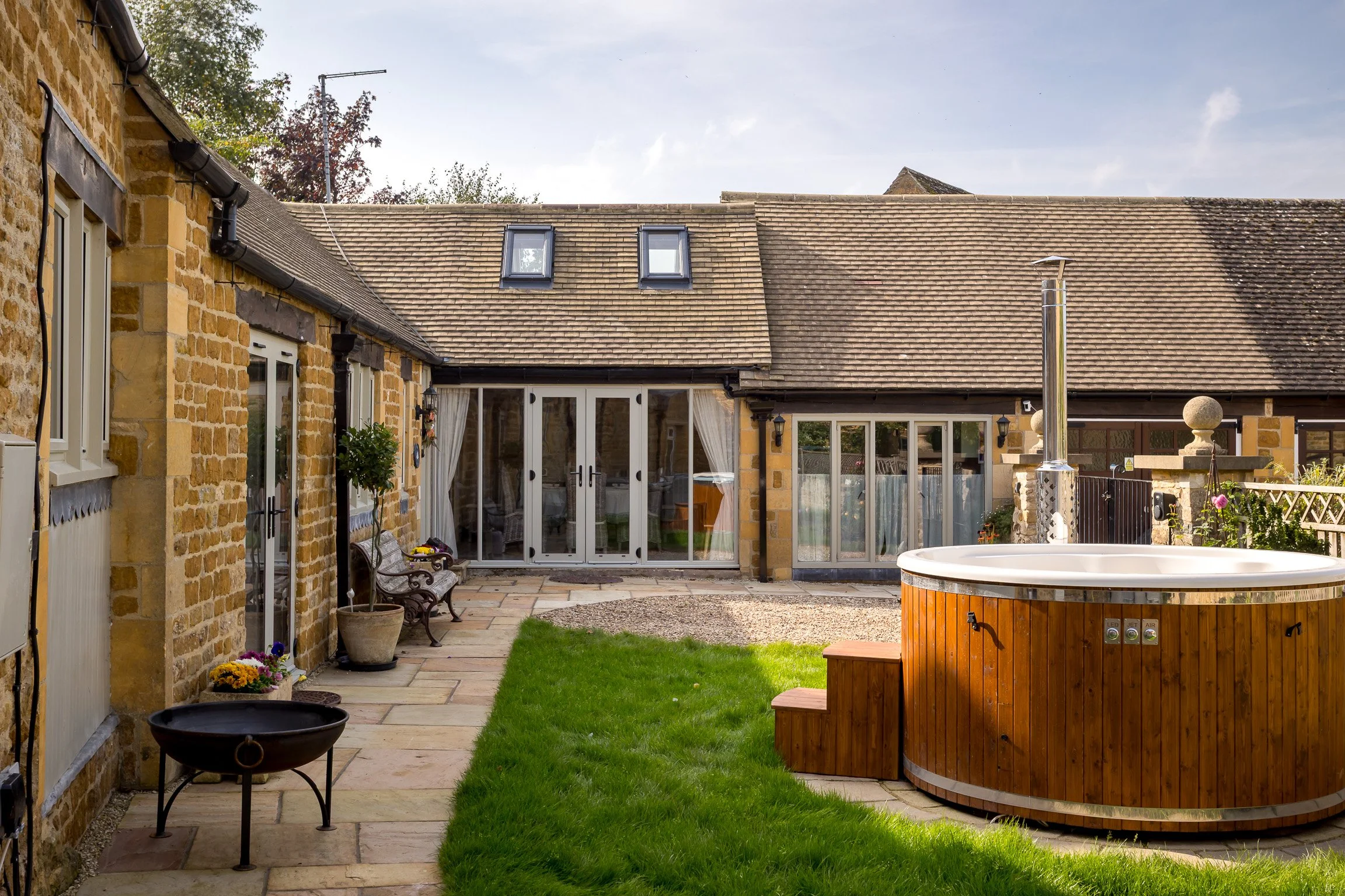 Backyard patio with stone paving, green grass, wooden hot tub, potted plants, outdoor seating with a bench, and glass sliding doors leading into a house with a stone exterior and a tiled roof.